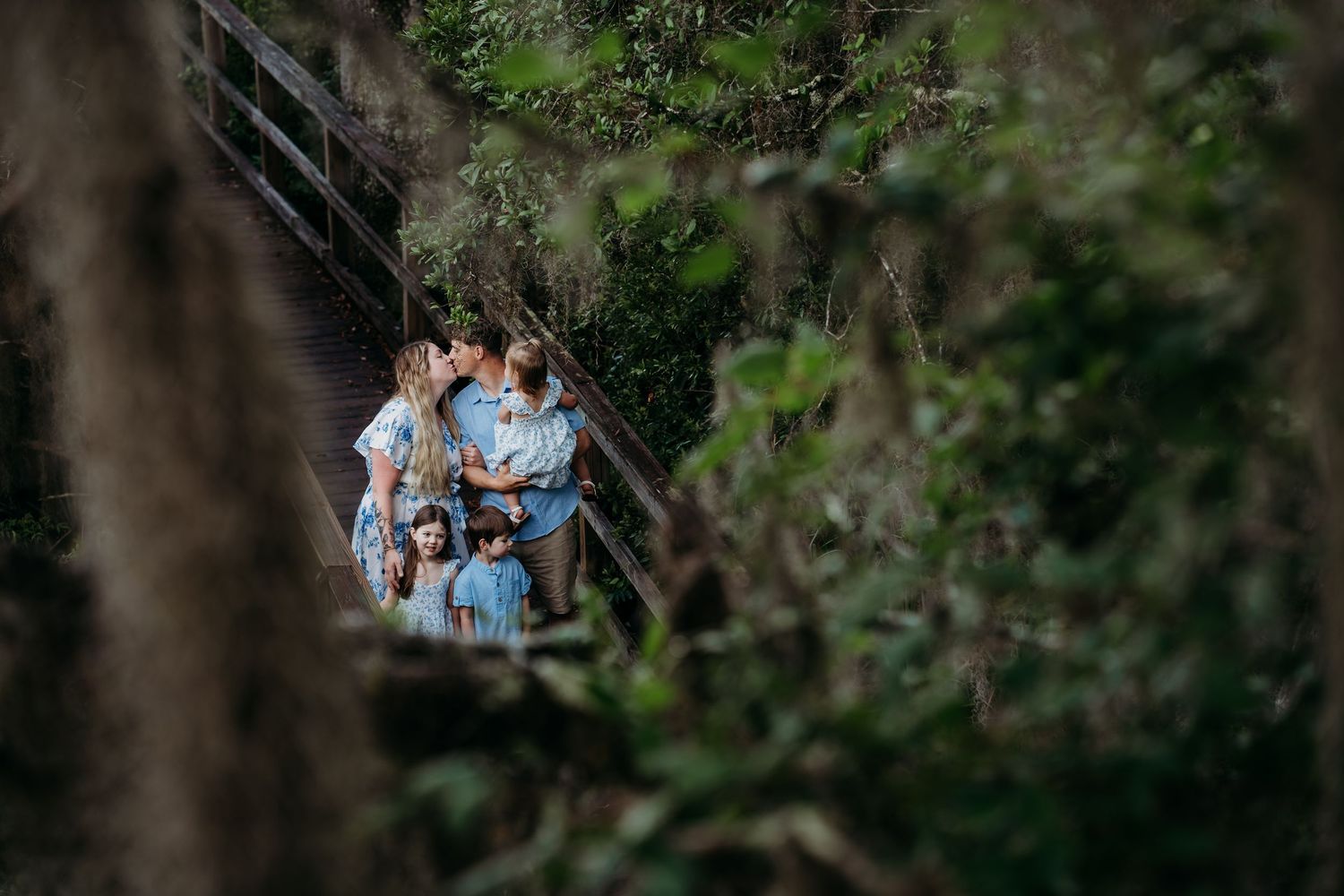 Family in matching blue outfits sharing a kiss on a wooden bridge surrounded by lush green trees.