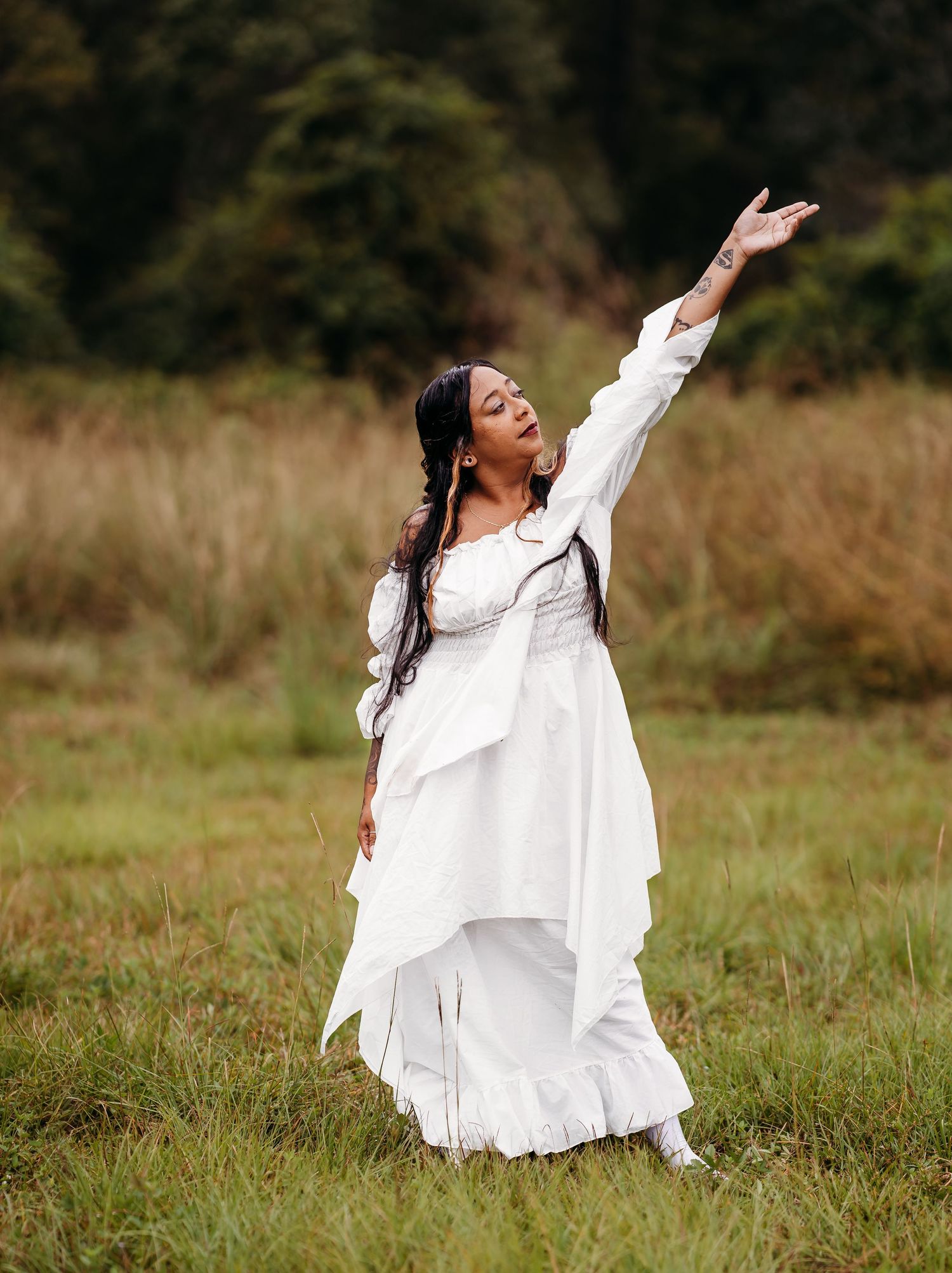 Woman in elegant white dress reaching arm skyward in open meadow with tall grass.
