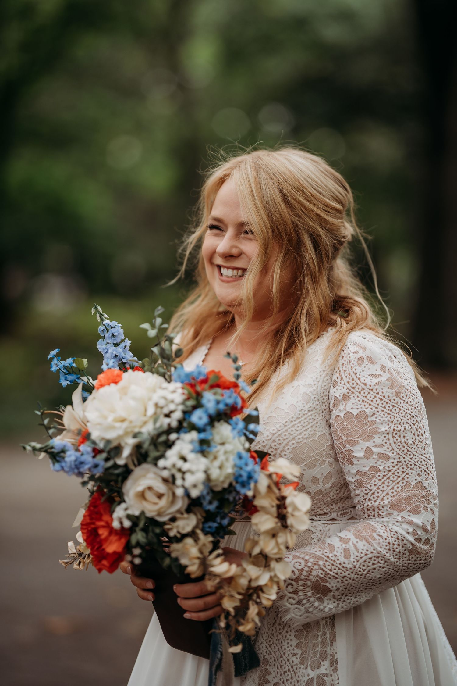 Smiling bride in lace gown holding vibrant red, white, and blue floral bouquet.