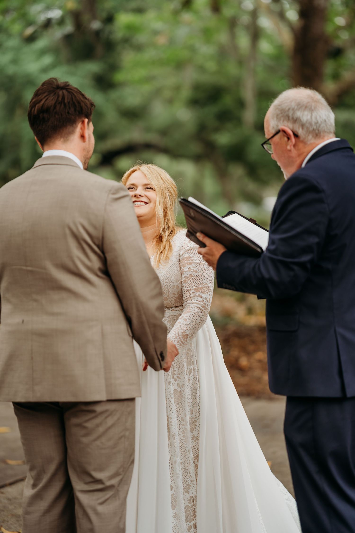 Smiling bride in lace gown holds grooms hands while officiant reads during outdoor wedding ceremony in park.