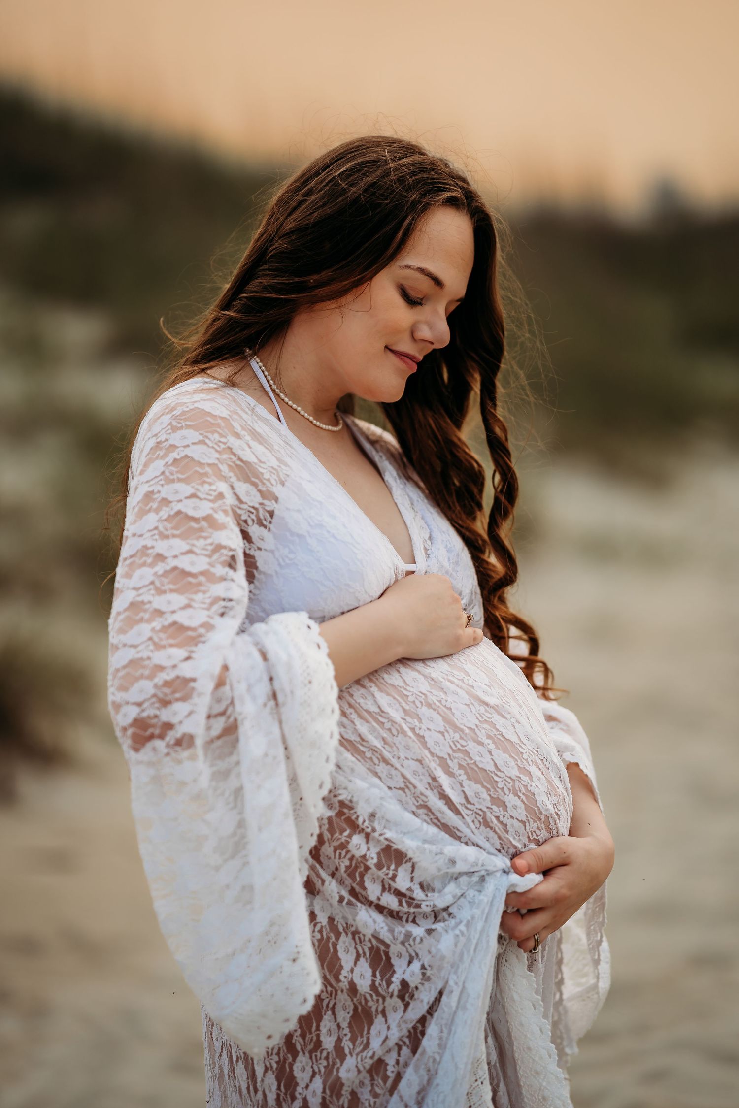 Pregnant woman in white lace dress cradles baby bump during golden hour beach maternity session.