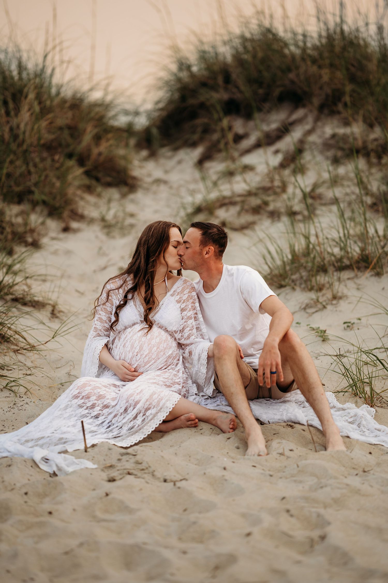 Couple shares a kiss while sitting together on sandy beach dunes during maternity photoshoot.