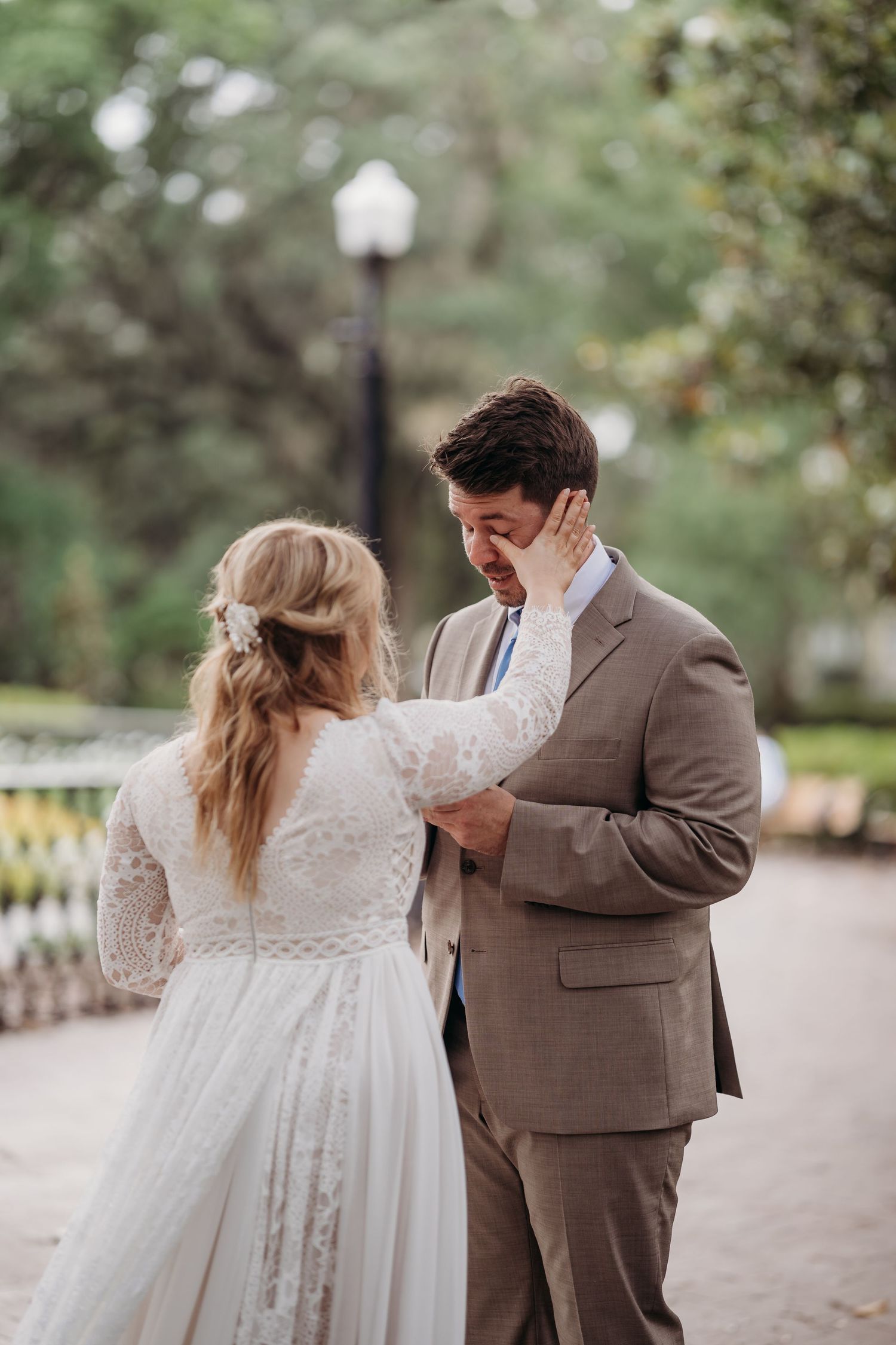 Bride in lace gown gently wipes tears from groom's cheek during heartfelt outdoor wedding ceremony.
