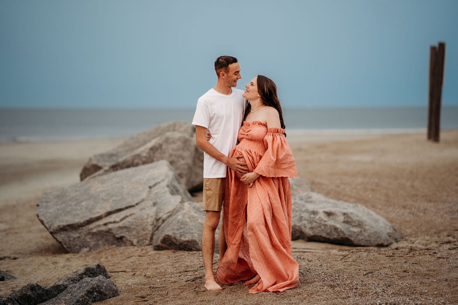 Expecting couple holding hands near rocky beach shore, woman in flowing coral maternity dress at sunset.