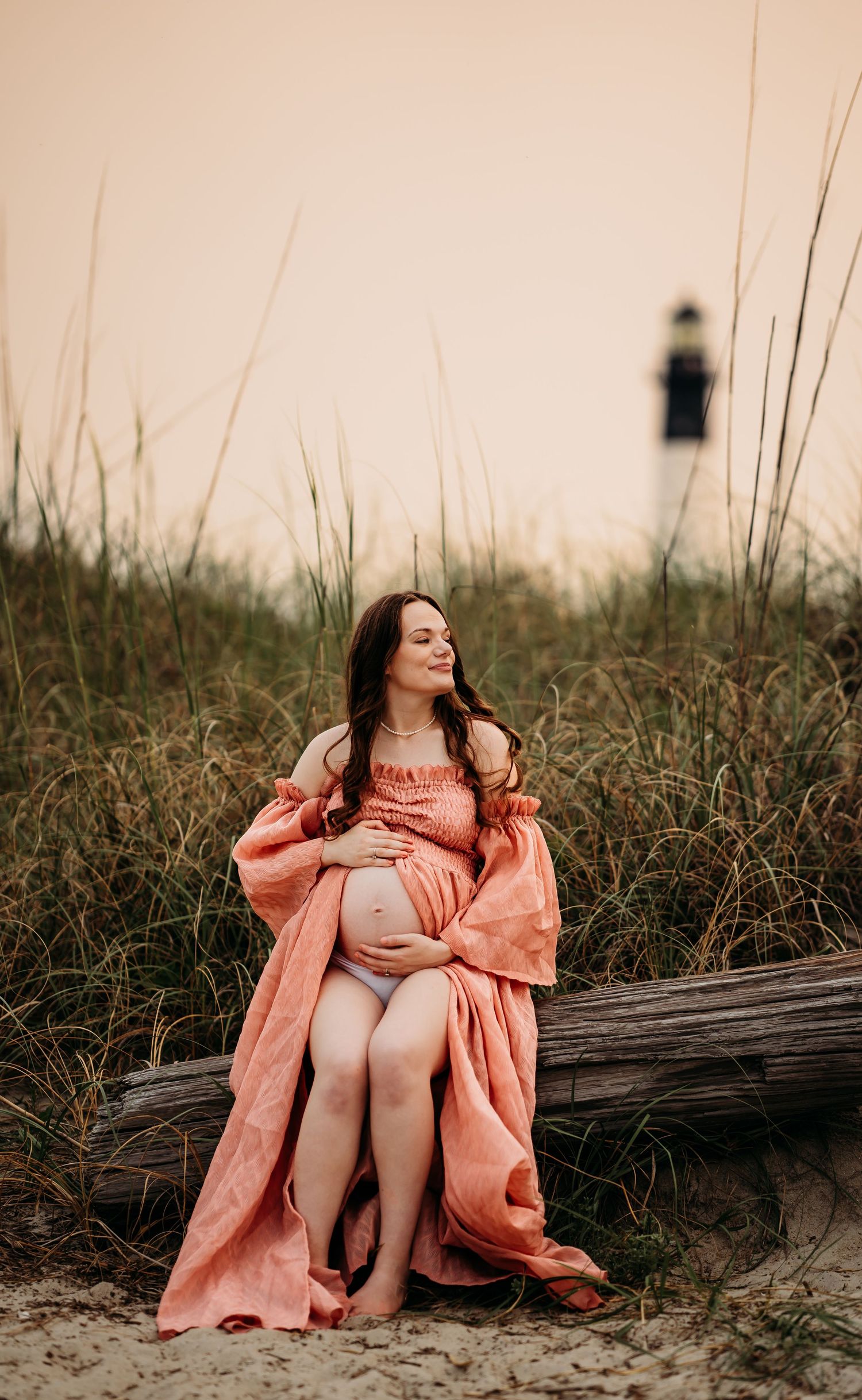 Pregnant woman in coral dress sitting on driftwood near lighthouse cradling baby bump at sunset.