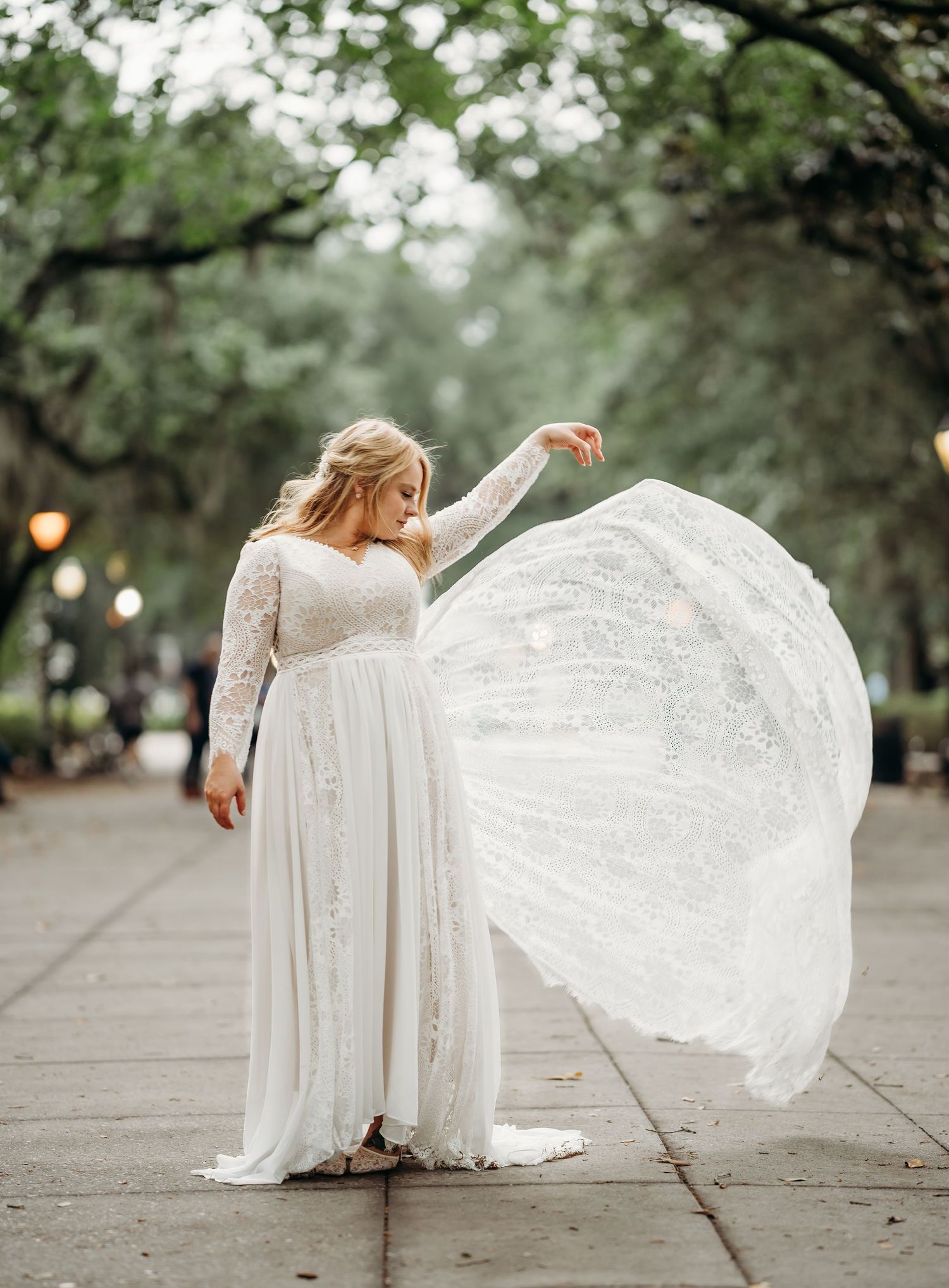 Bride twirls flowing lace wedding dress on tree-lined park pathway at dusk.