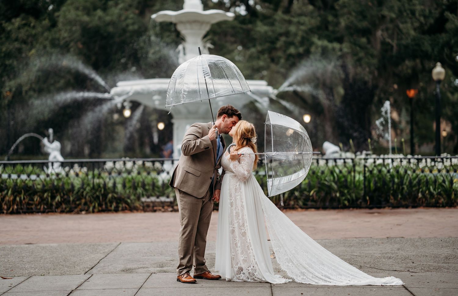 Bride and groom kiss under clear umbrella in front of ornate fountain.