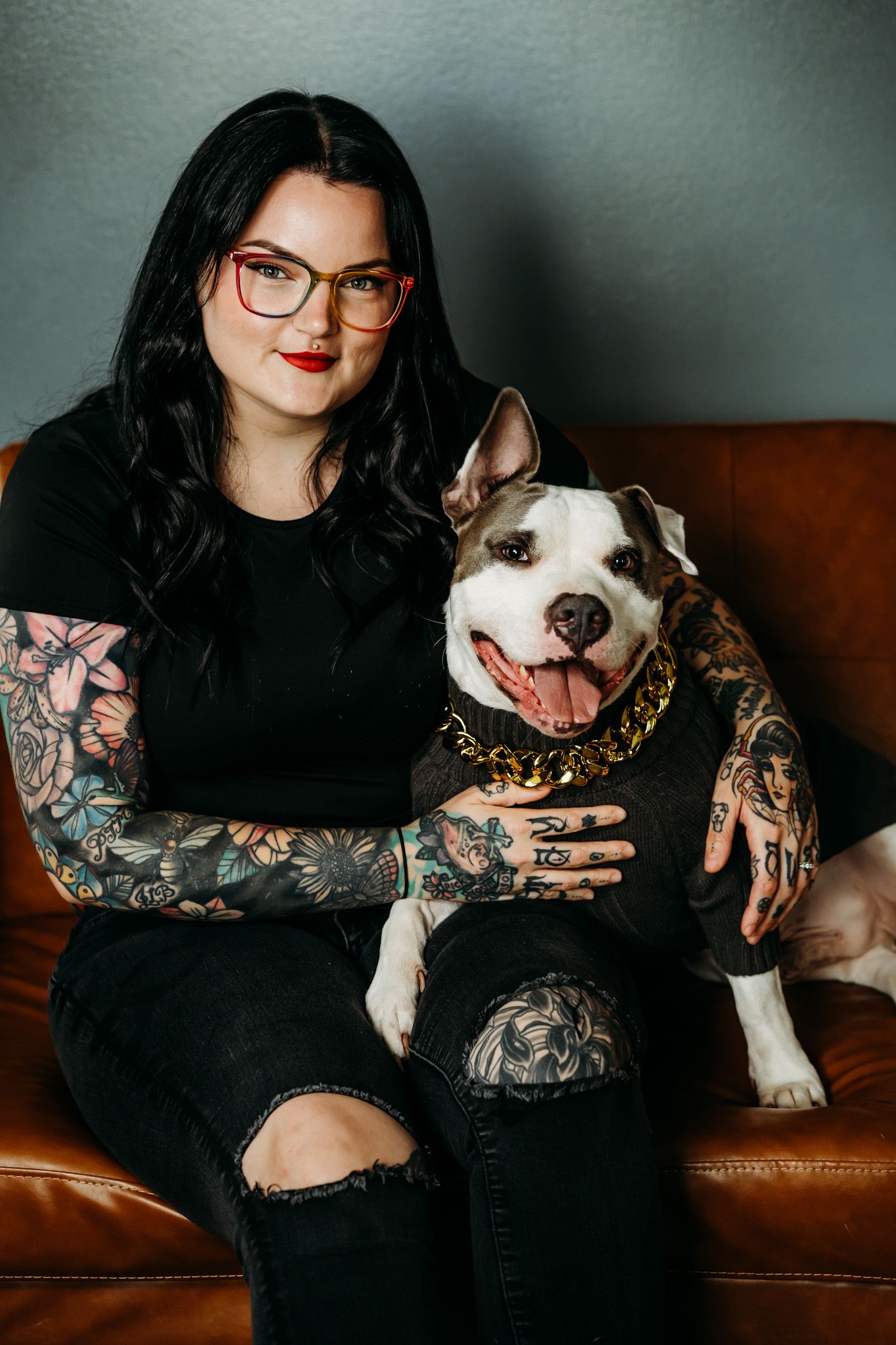 Tattooed woman with red glasses sitting on leather couch with smiling pit bull dog.
