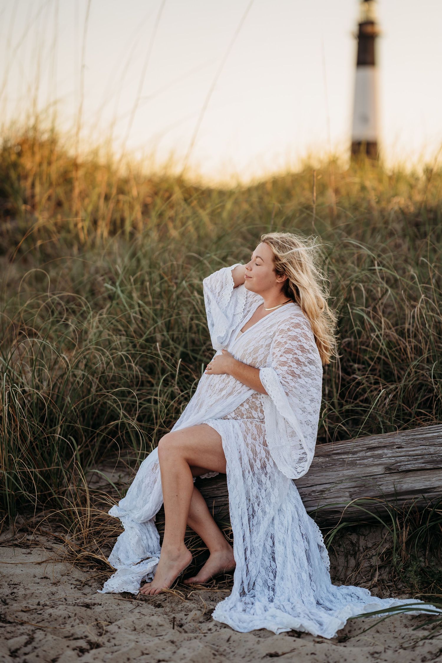Pregnant blonde woman in white lace gown sits on driftwood at golden hour beach near lighthouse.