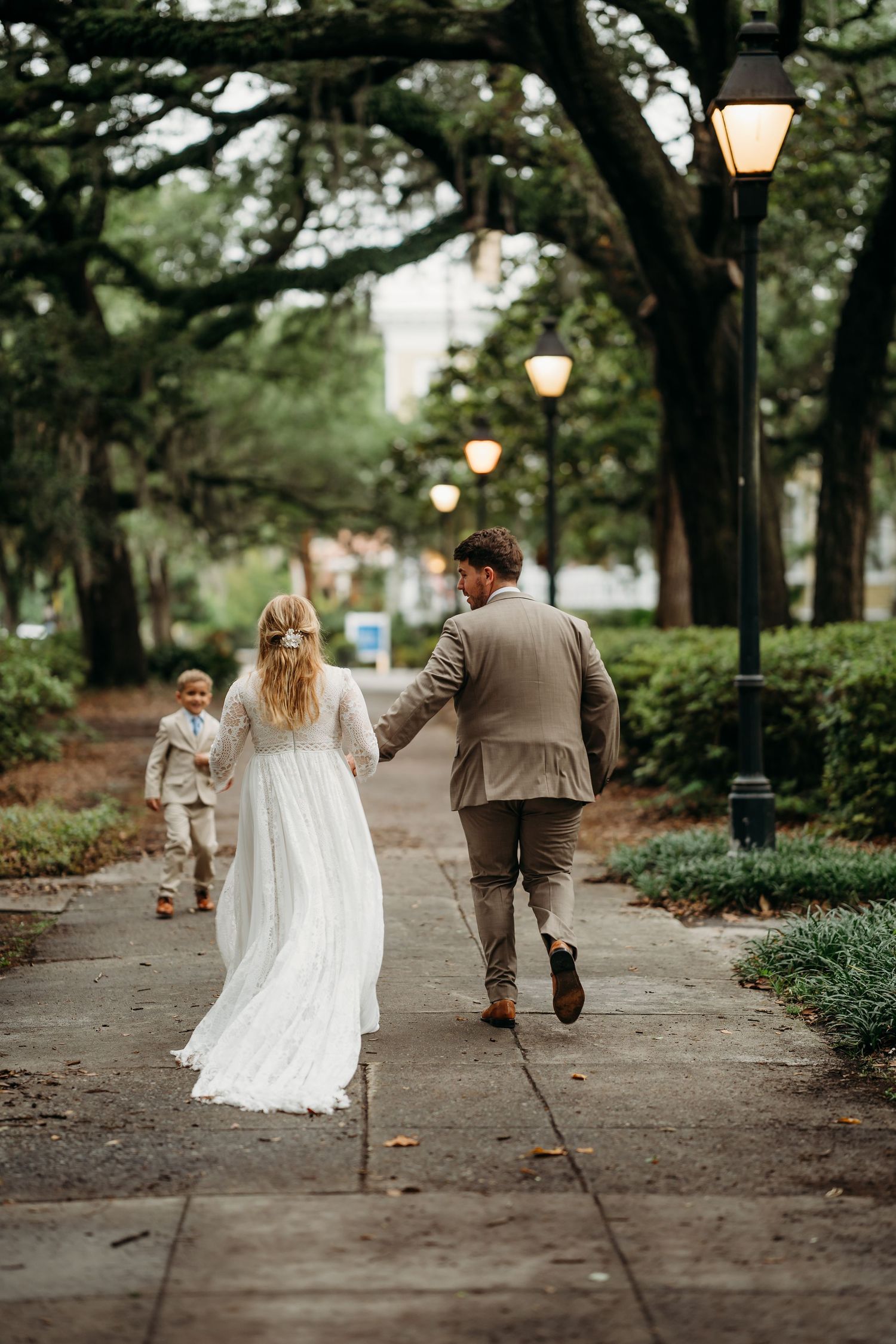 Bride and groom walk hand-in-hand with young boy on lamp-lit park pathway.