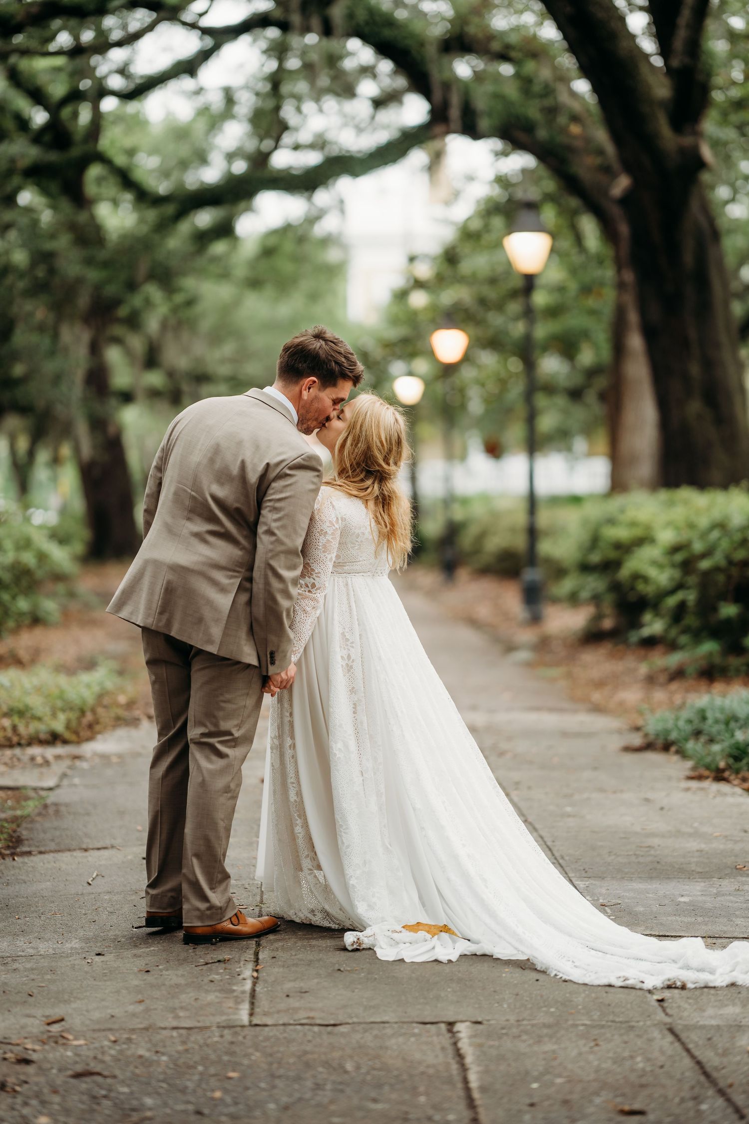 Groom in tan suit leans to kiss bride in elegant white gown on a romantic lamp-lit tree-lined garden walkway.