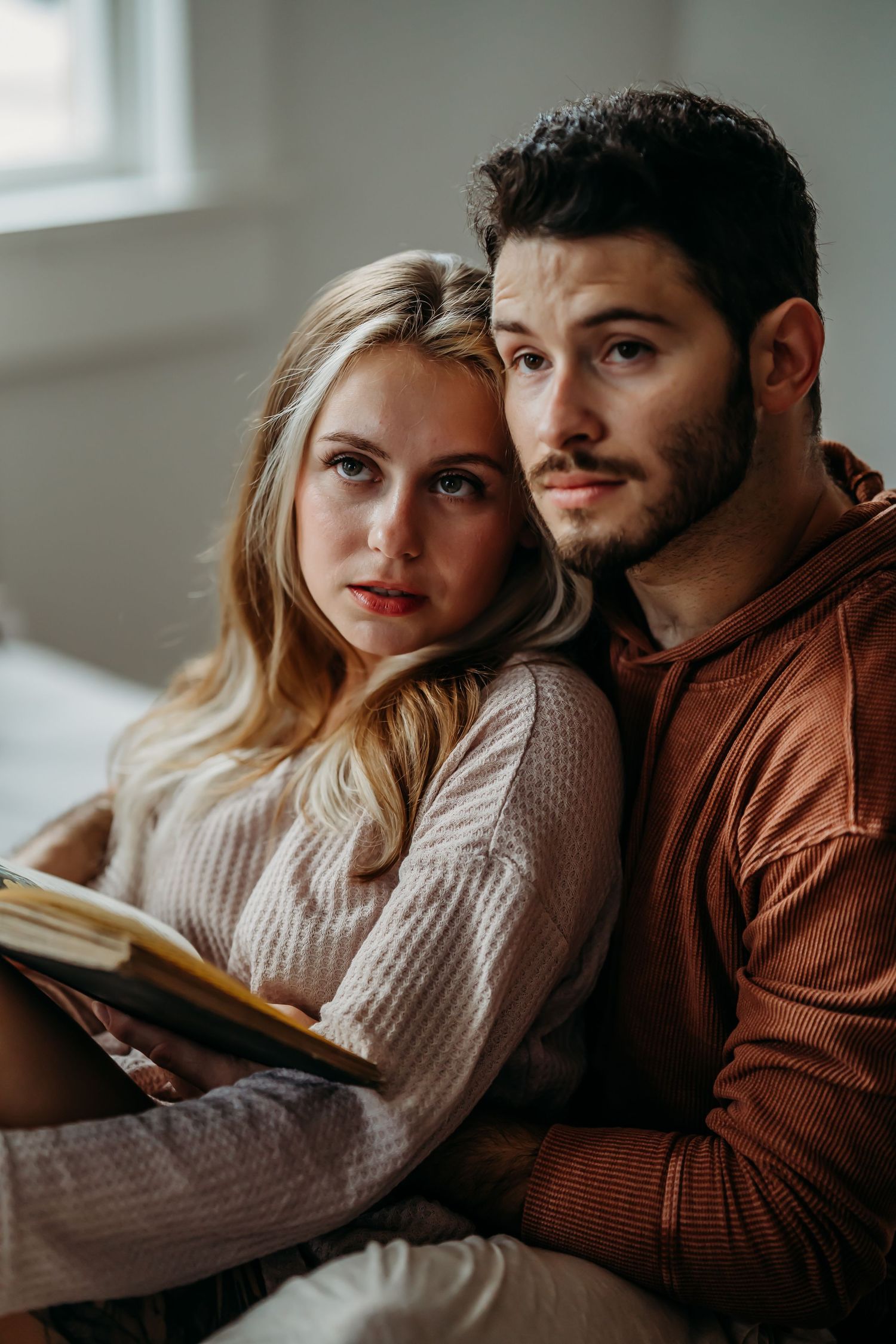 A couple leans together holding a book, posing for a portrait in a softly lit bedroom.