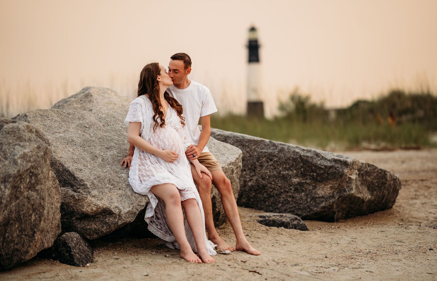 Couple sharing a tender moment on beach rocks near a lighthouse during a maternity photo session.