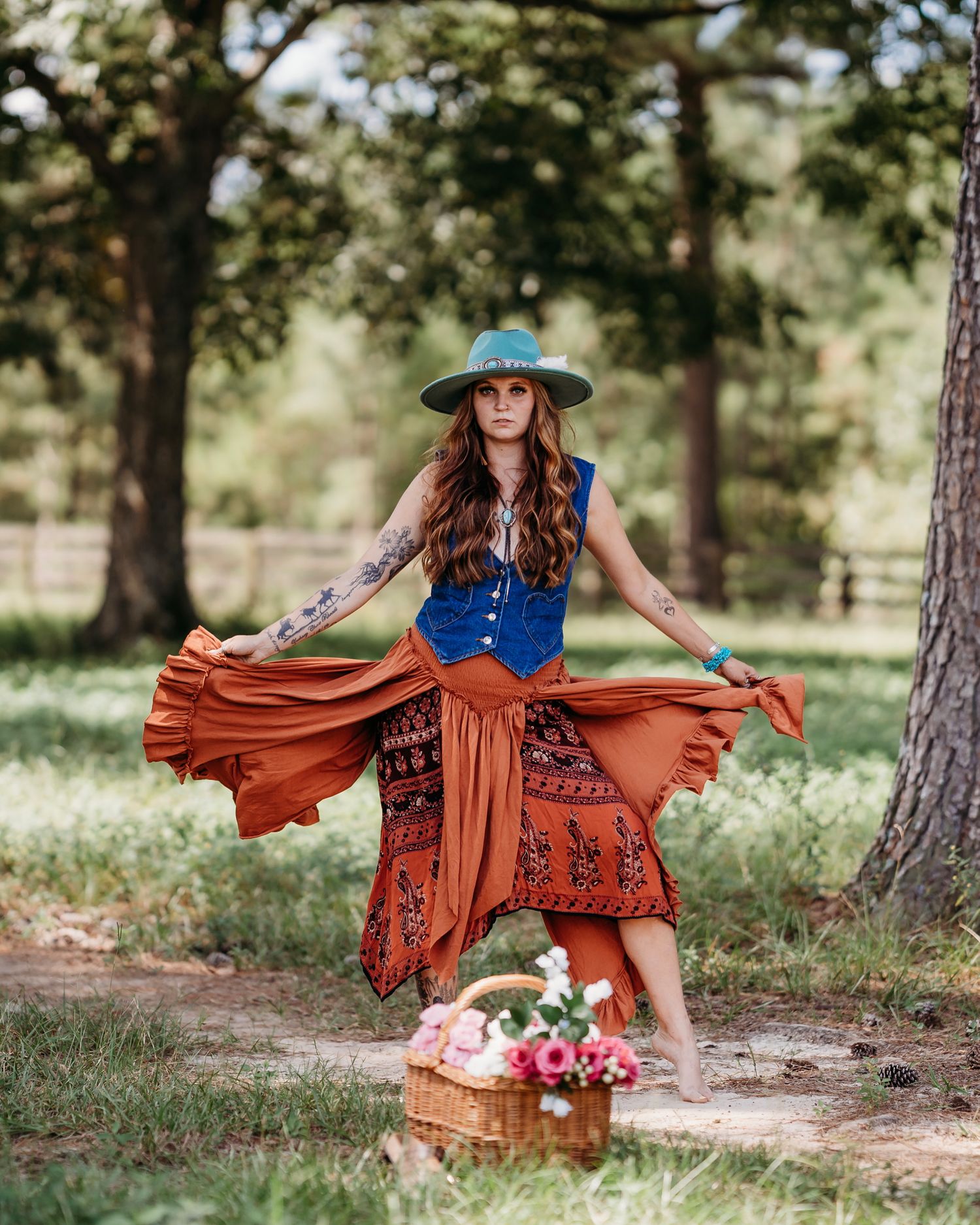 Bohemian woman in blue vest, rust skirt, and teal hat poses outdoors near a floral basket in a wooded setting.