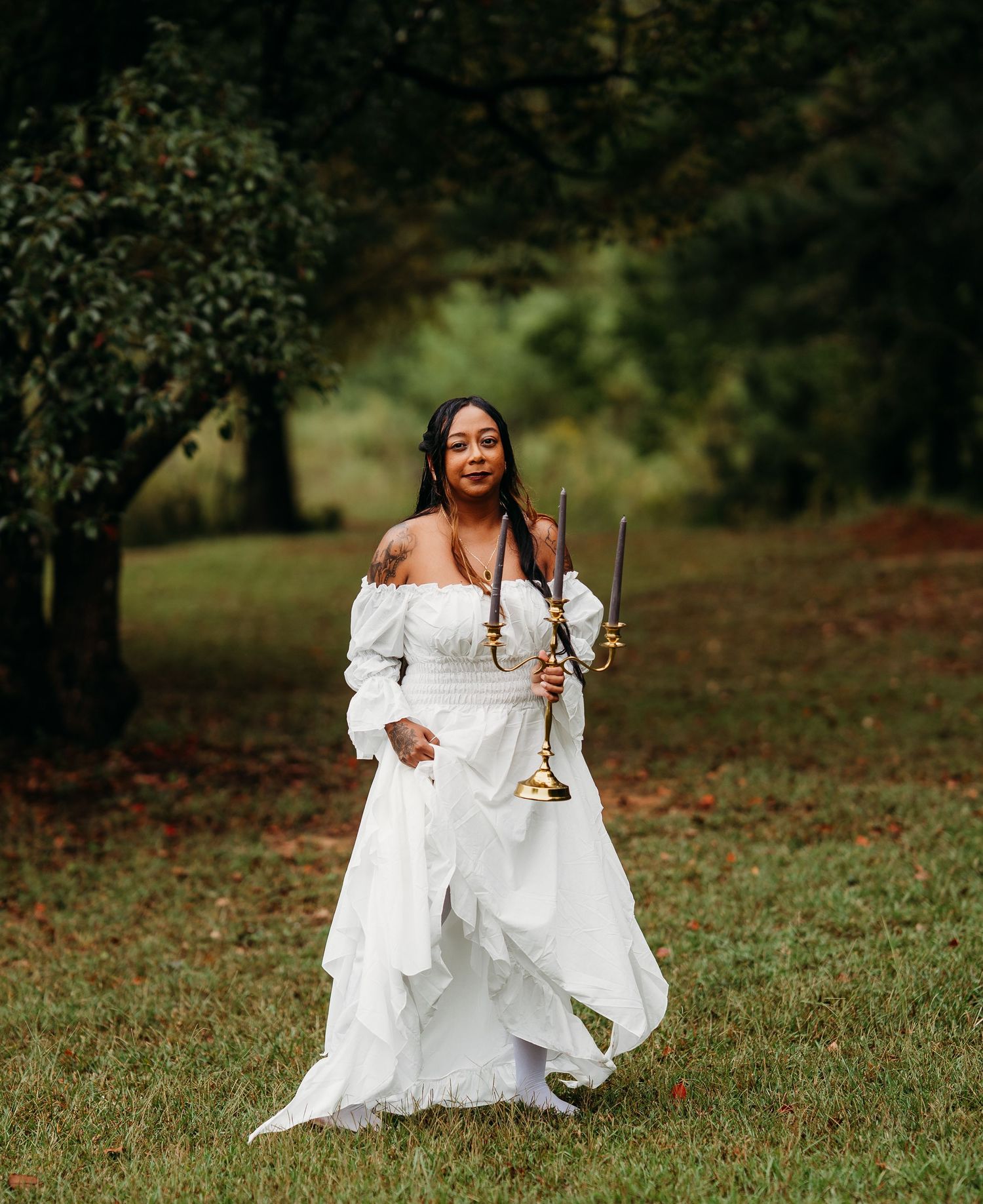 Woman in white off-shoulder gown walks through park carrying gold candelabra with dark candles outdoors.