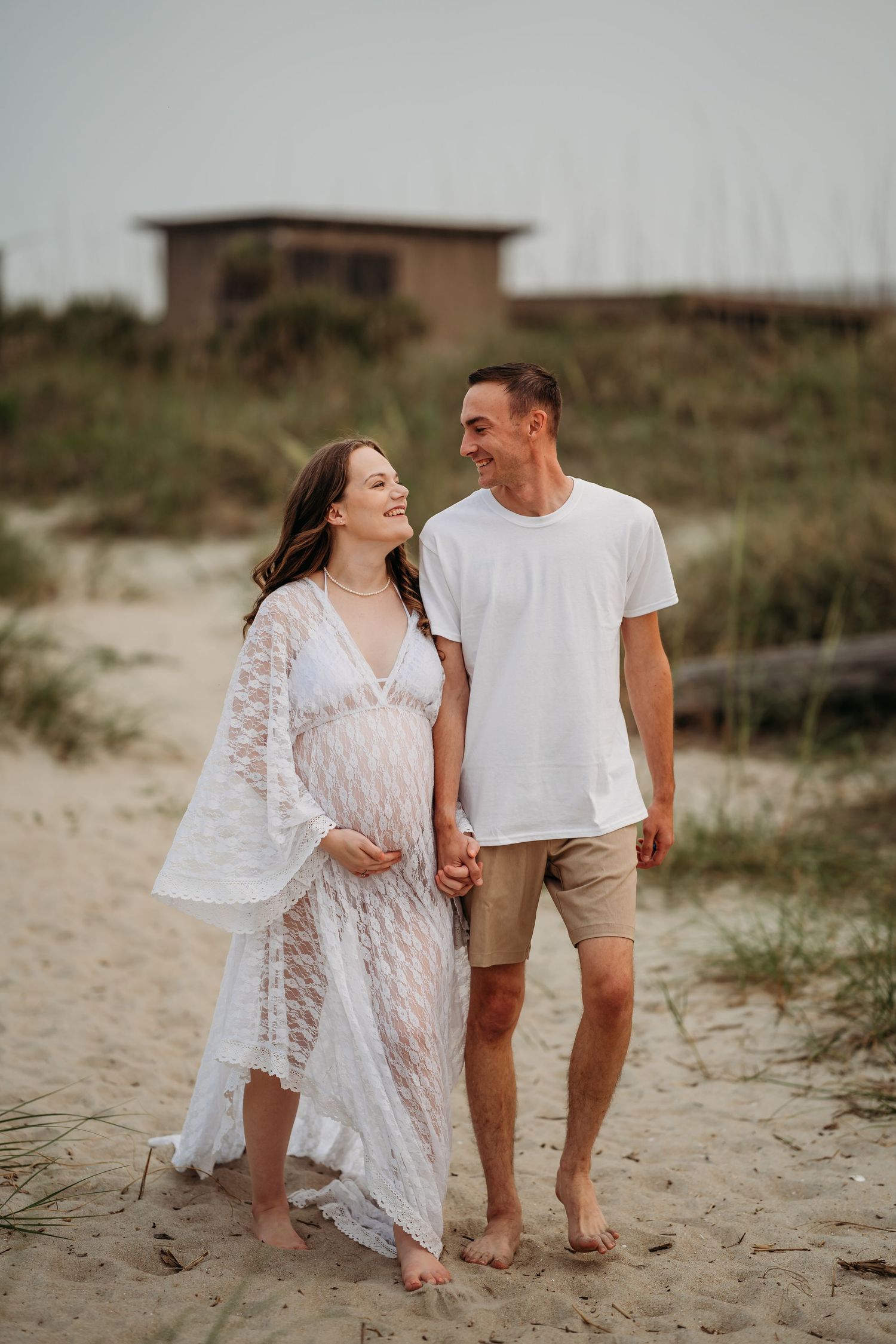 Happy expecting couple walking hand in hand on sandy beach, laughing together at sunset.