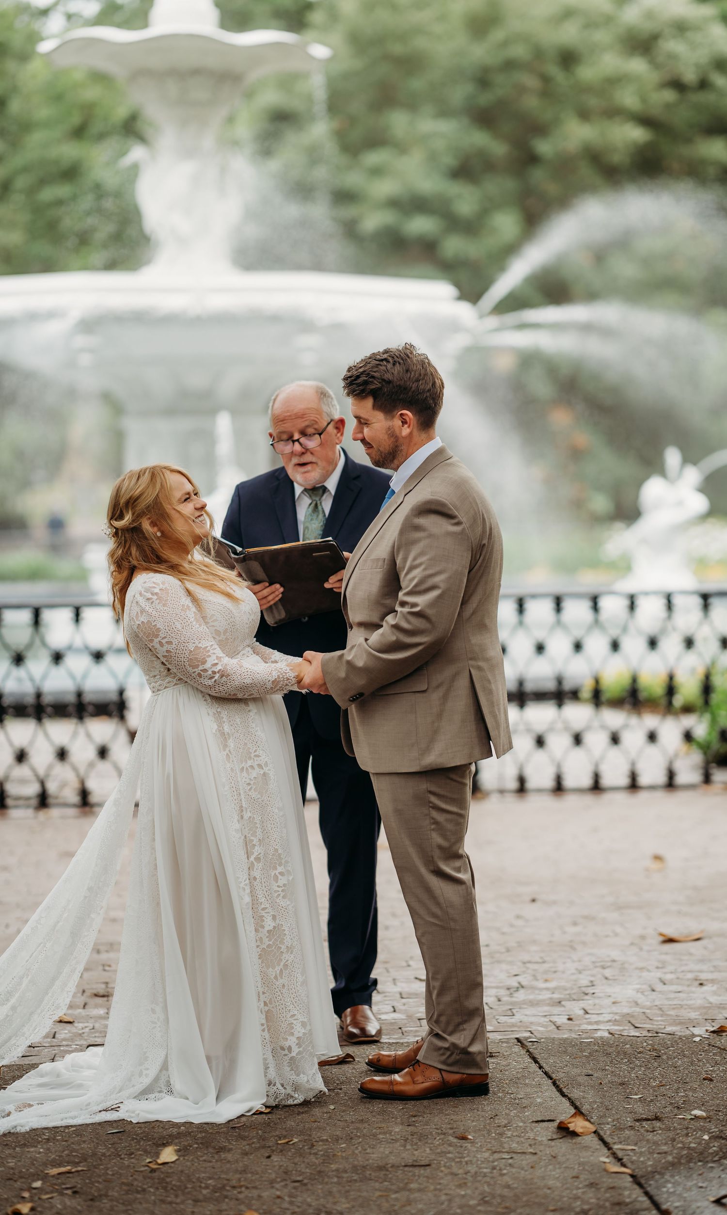 Bride in lace gown and groom in tan suit hold hands during vows at Forsyth Park fountain ceremony.