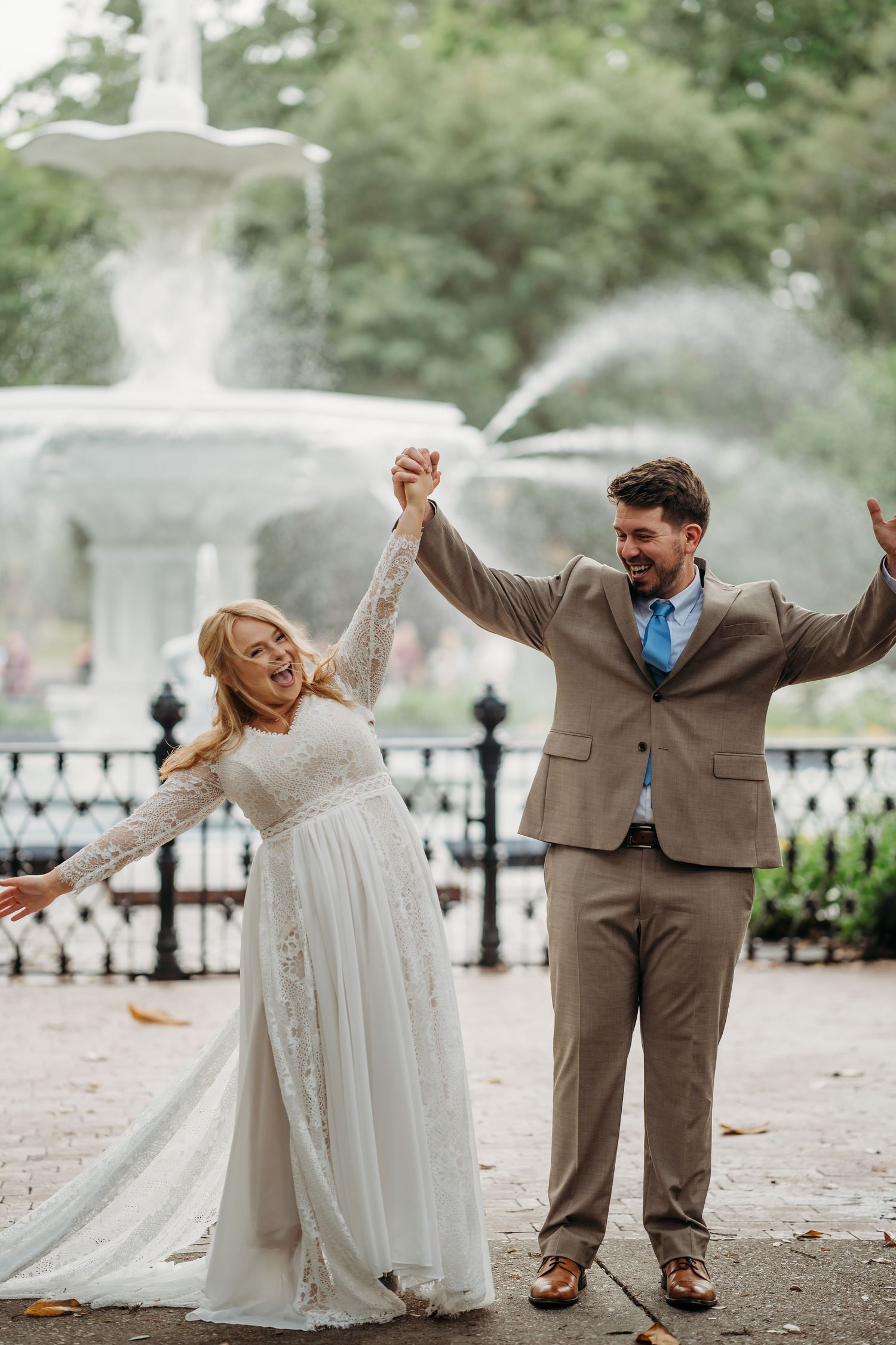Jubilant bride and groom raise their hands in celebration in front of a large ornate fountain.