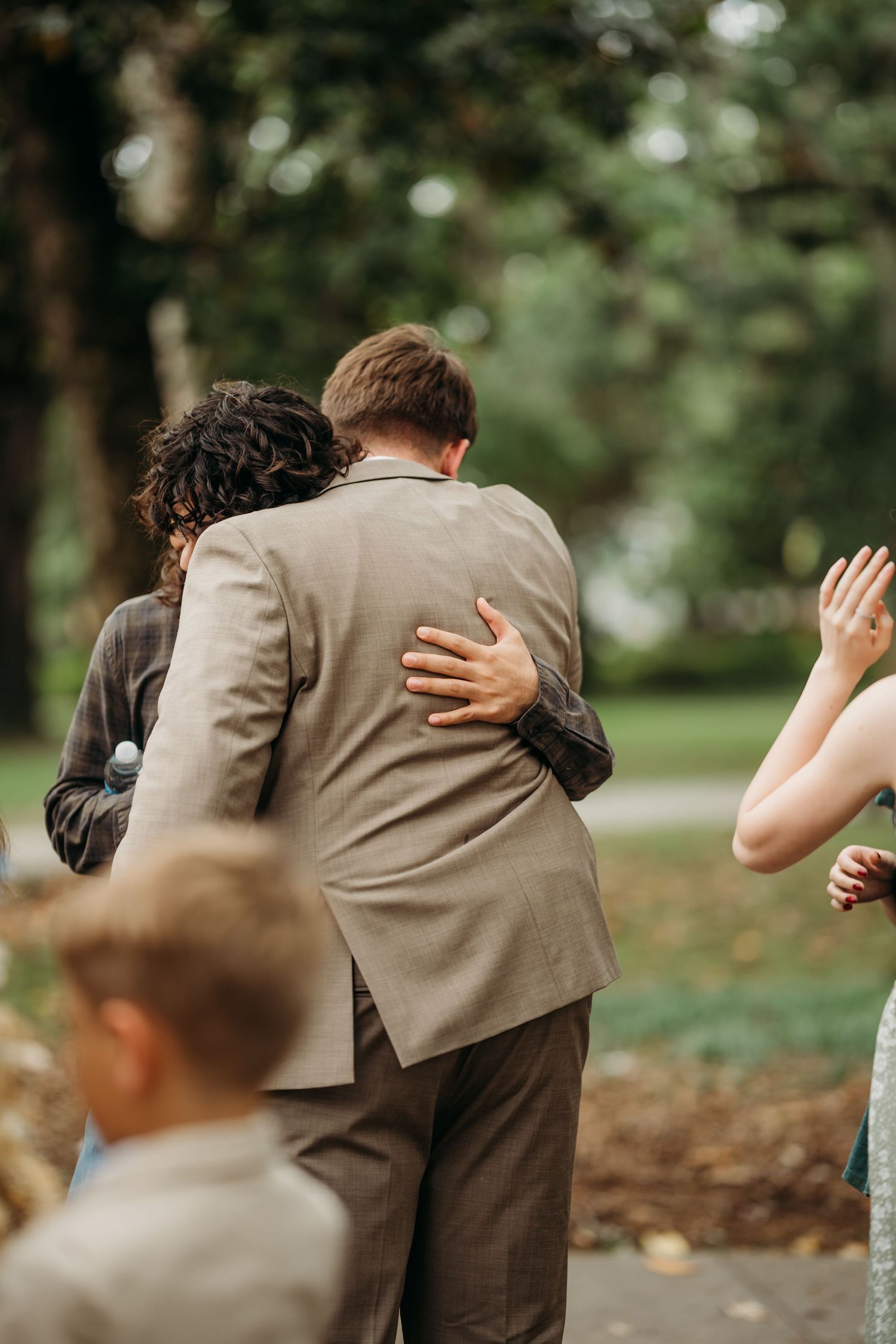 Wedding guest embraces groom in tan suit from behind in a tree-lined park setting.