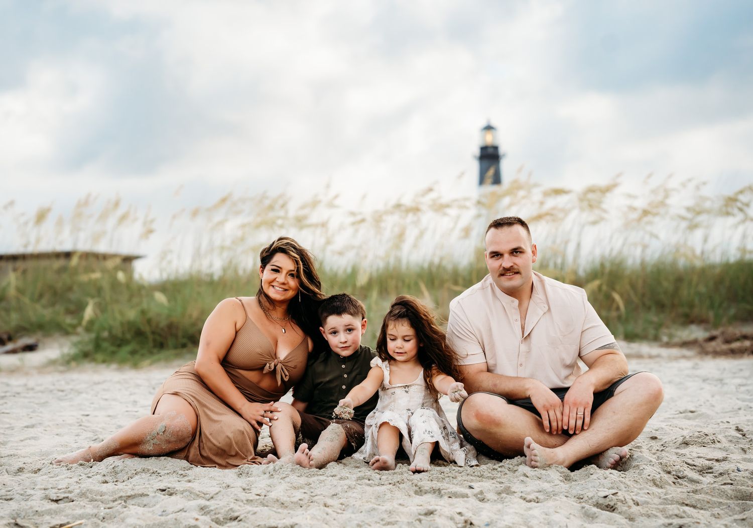 Family of four sitting together on sandy beach with lighthouse and sea oats in background.