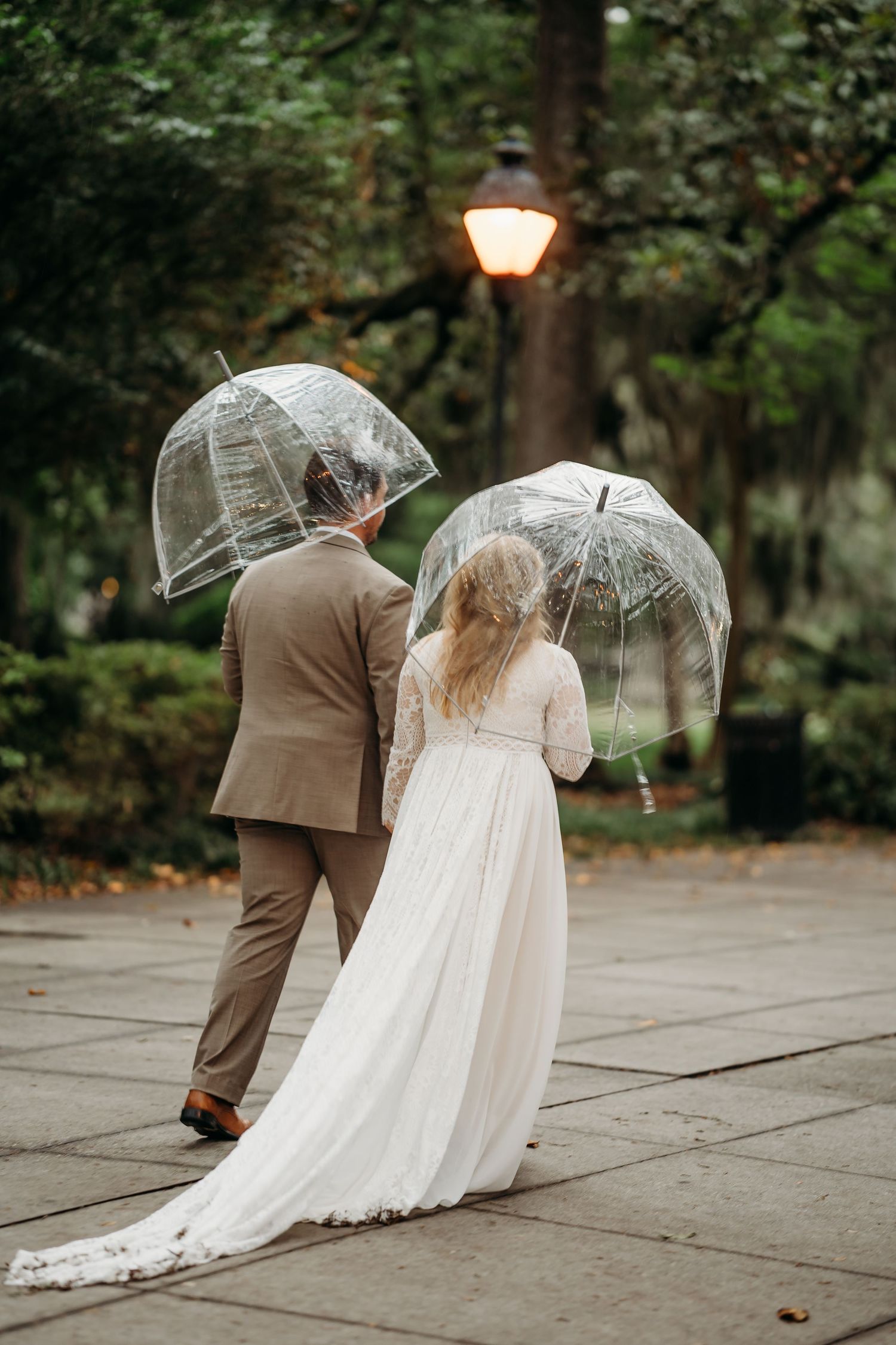 Bride and groom walking away holding clear bubble umbrellas on a rainy wedding day in a park.