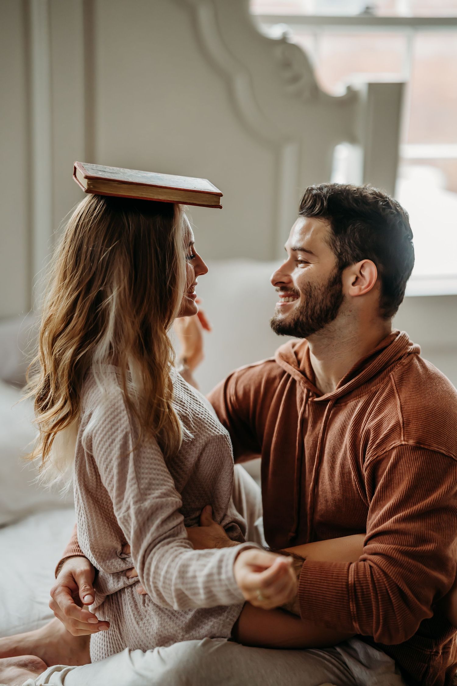 Happy couple sitting on bed, woman balancing book on head, smiling at bearded man in brown hoodie.