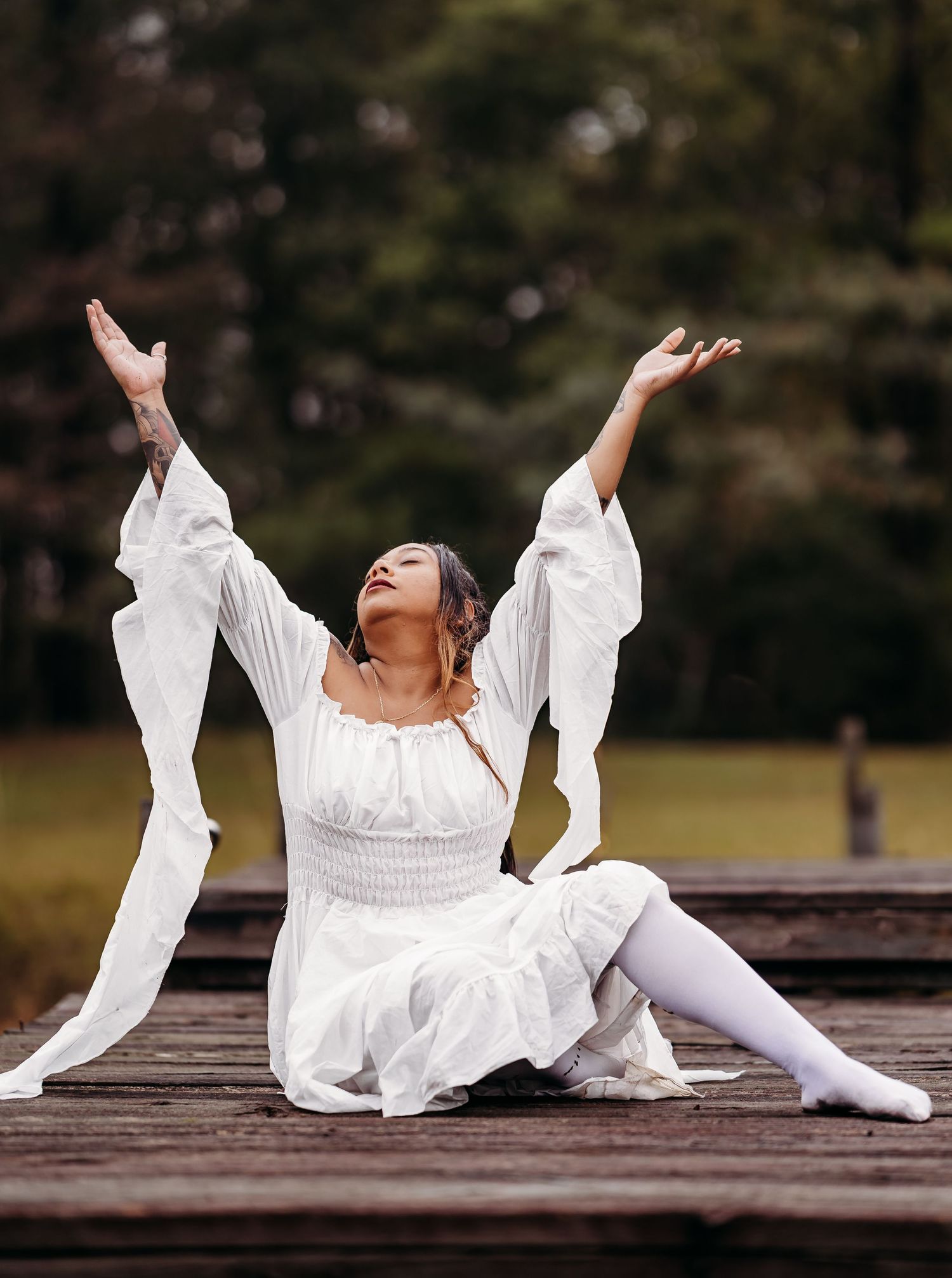 Woman in white dress sits on a dock with both arms raised joyfully toward the sky.
