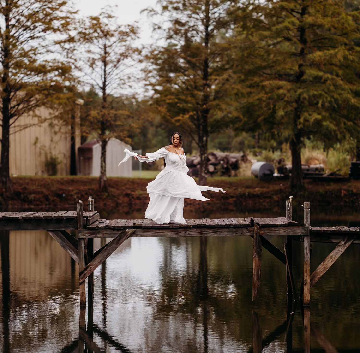 Bride in flowing white wedding dress twirling on wooden dock over reflective pond surrounded by autumn trees.