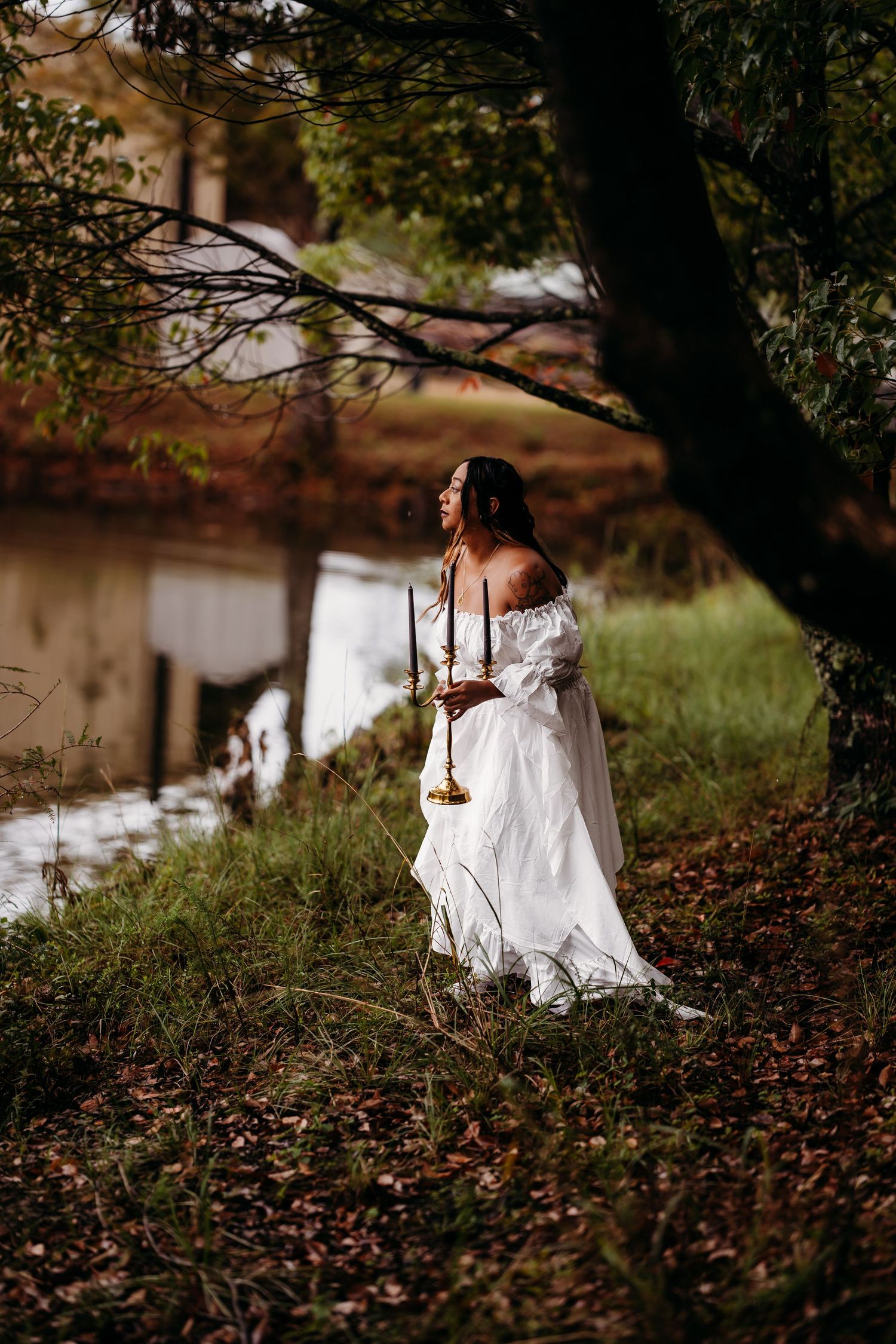 Bride in flowing white dress holding candles near serene woodland pond.