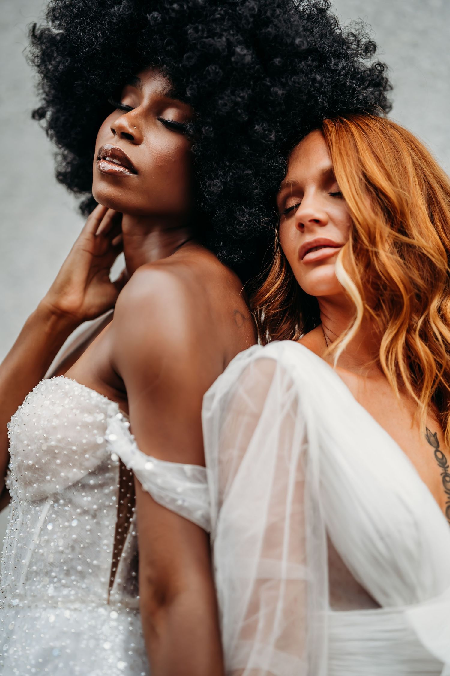 Two elegant brides in white dresses pose together, one with an afro and one with auburn waves.