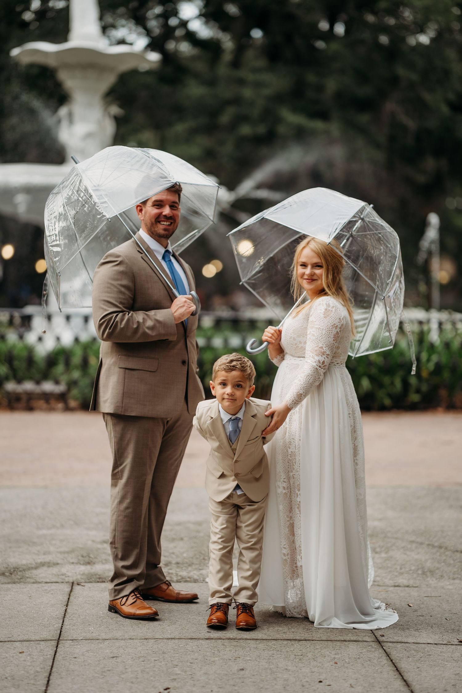 Wedding couple with young boy in matching suit pose under clear bubble umbrellas before white fountain.