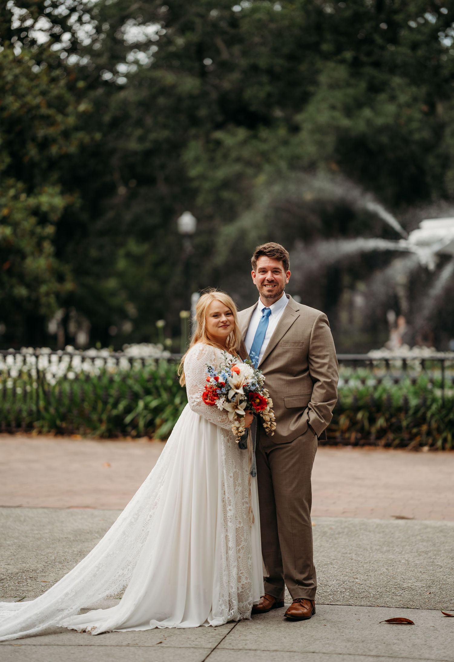 Bride and groom smile at camera in park setting with white fountain and trees behind them.