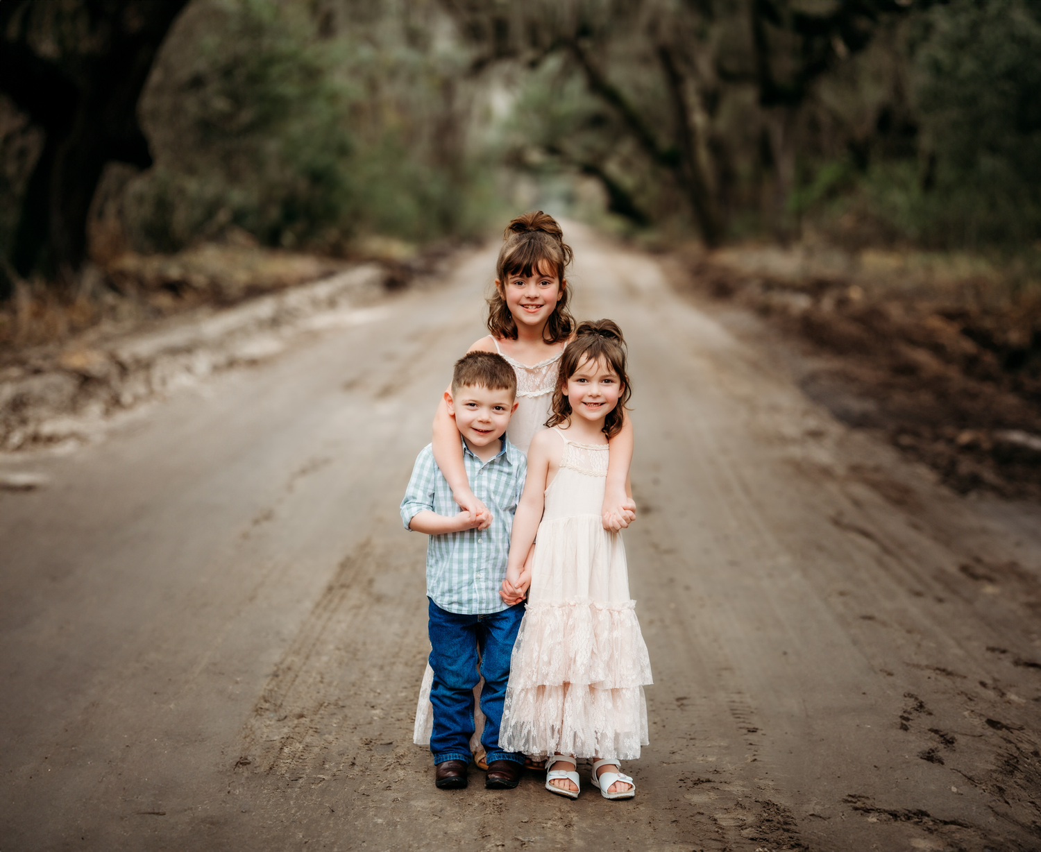 Three young children pose together on a scenic dirt road surrounded by mossy oak trees.
