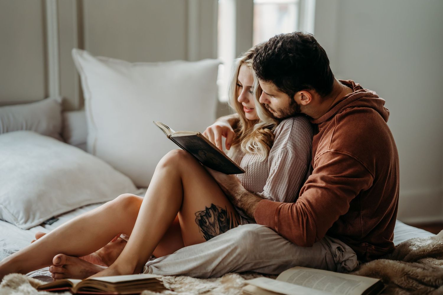 A romantic couple reads a book together while relaxing on a cozy bed with soft natural lighting.