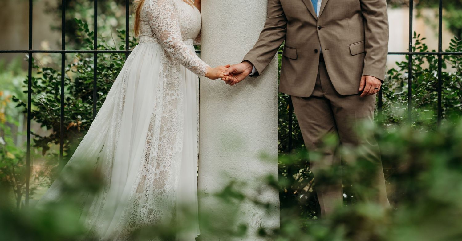 Close-up of bride and groom holding hands in wedding attire, bride in lace gown and groom in tan suit.
