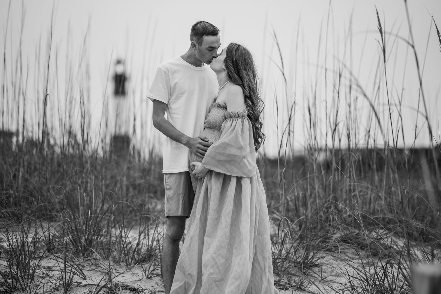 Black and white photo of pregnant couple sharing a tender moment near beach dunes and lighthouse.