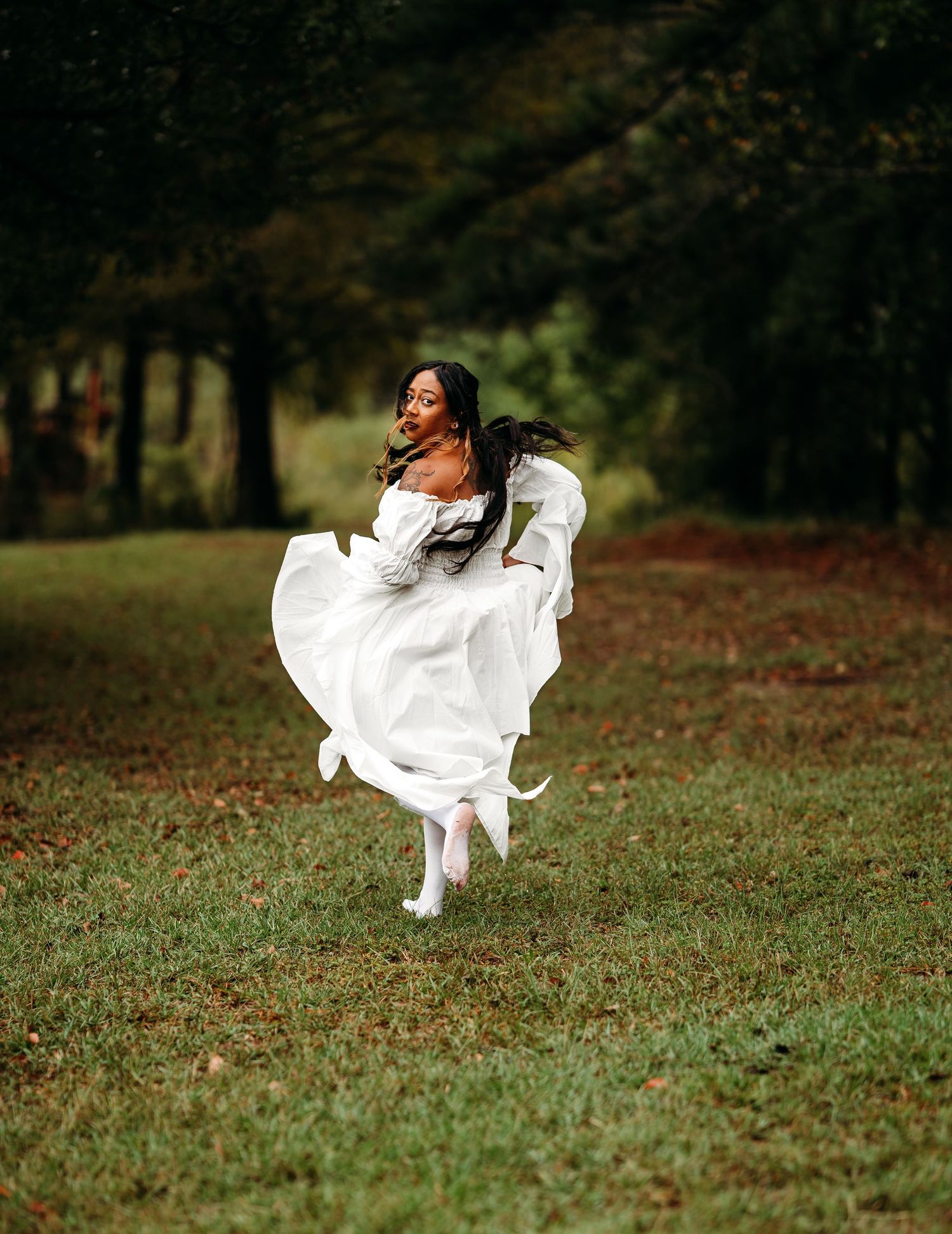 Woman in white off-shoulder dress runs through grassy field with trees in background on overcast day.