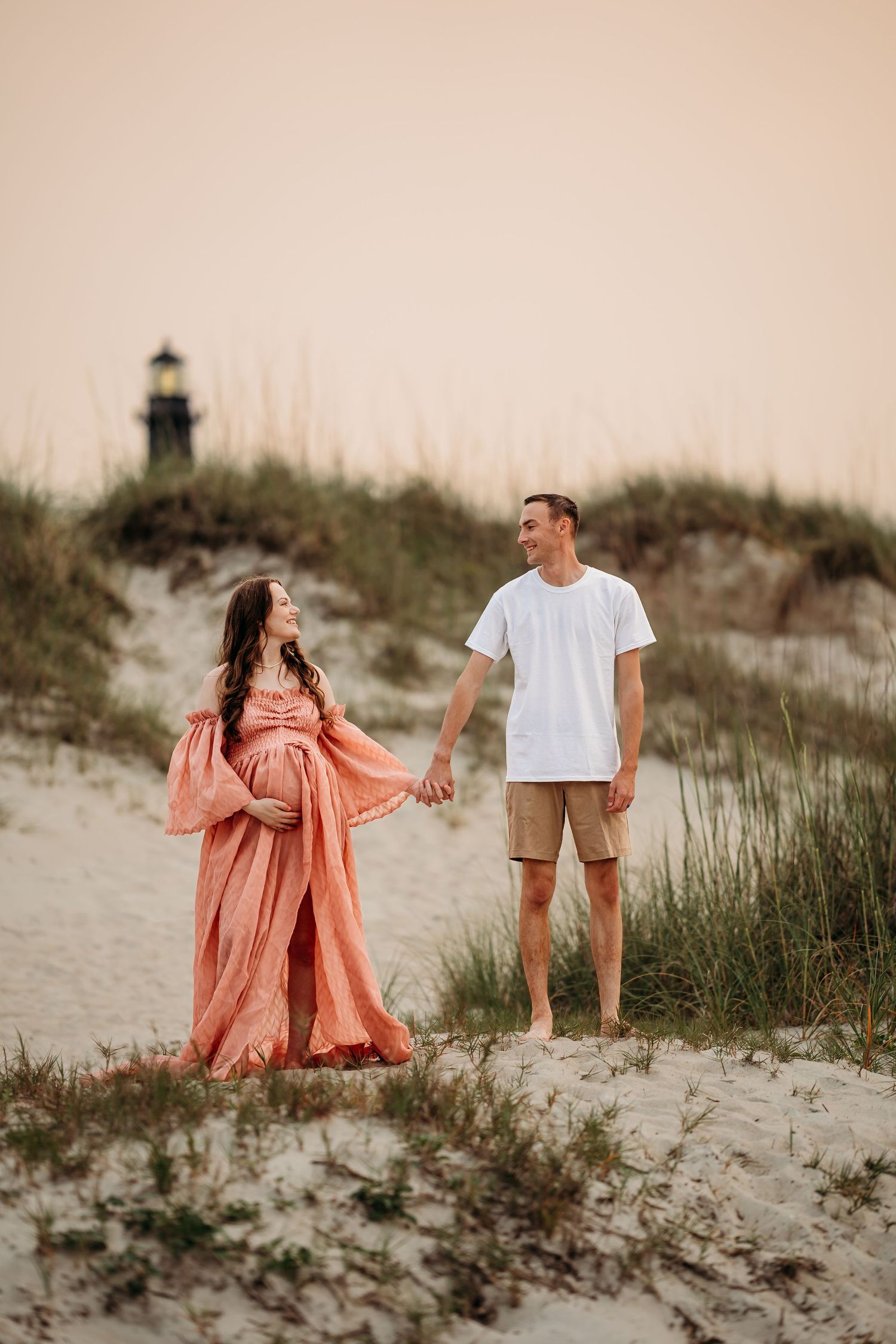 Pregnant couple holding hands on sandy beach dunes at sunset with lighthouse visible in background.