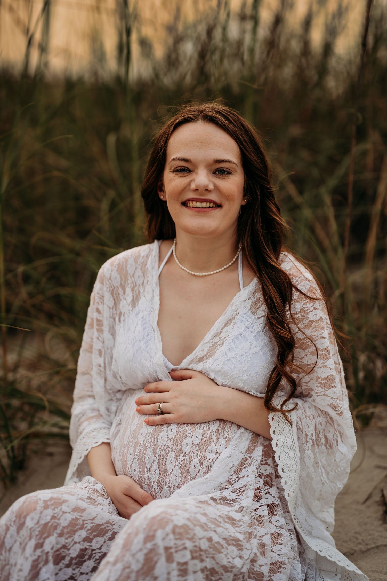 Smiling pregnant woman in white lace maternity gown cradling her baby bump at golden hour beach.