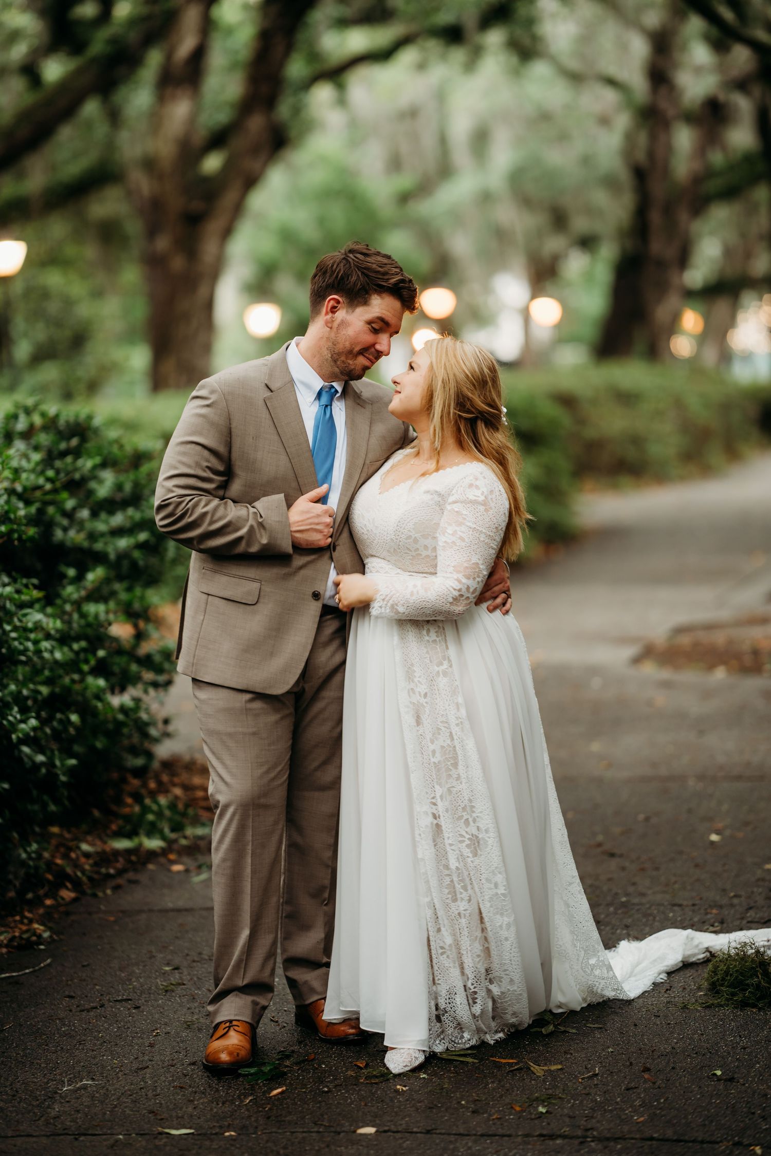 Bride and groom gazing lovingly at each other on a romantic tree-lined pathway with glowing lights behind them.