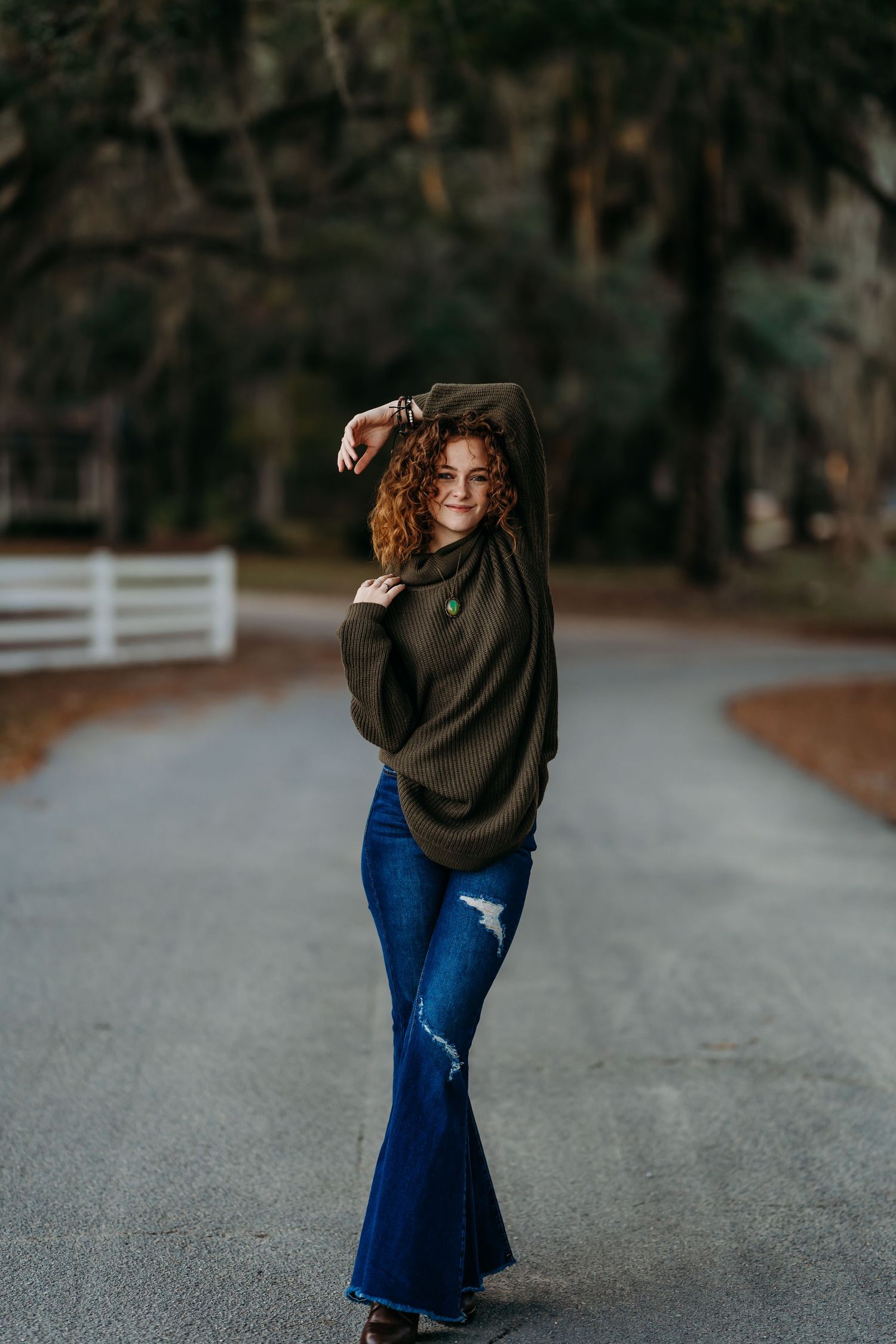 Curly redhead teen poses confidently on winding road in flare jeans for senior portraits.