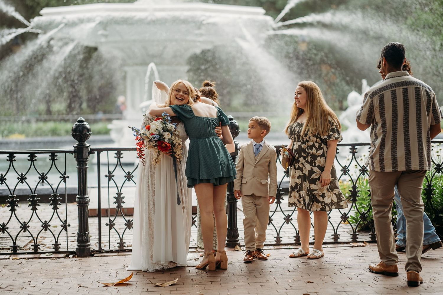 Bride embraces a guest in a teal dress near a grand fountain surrounded by family members.