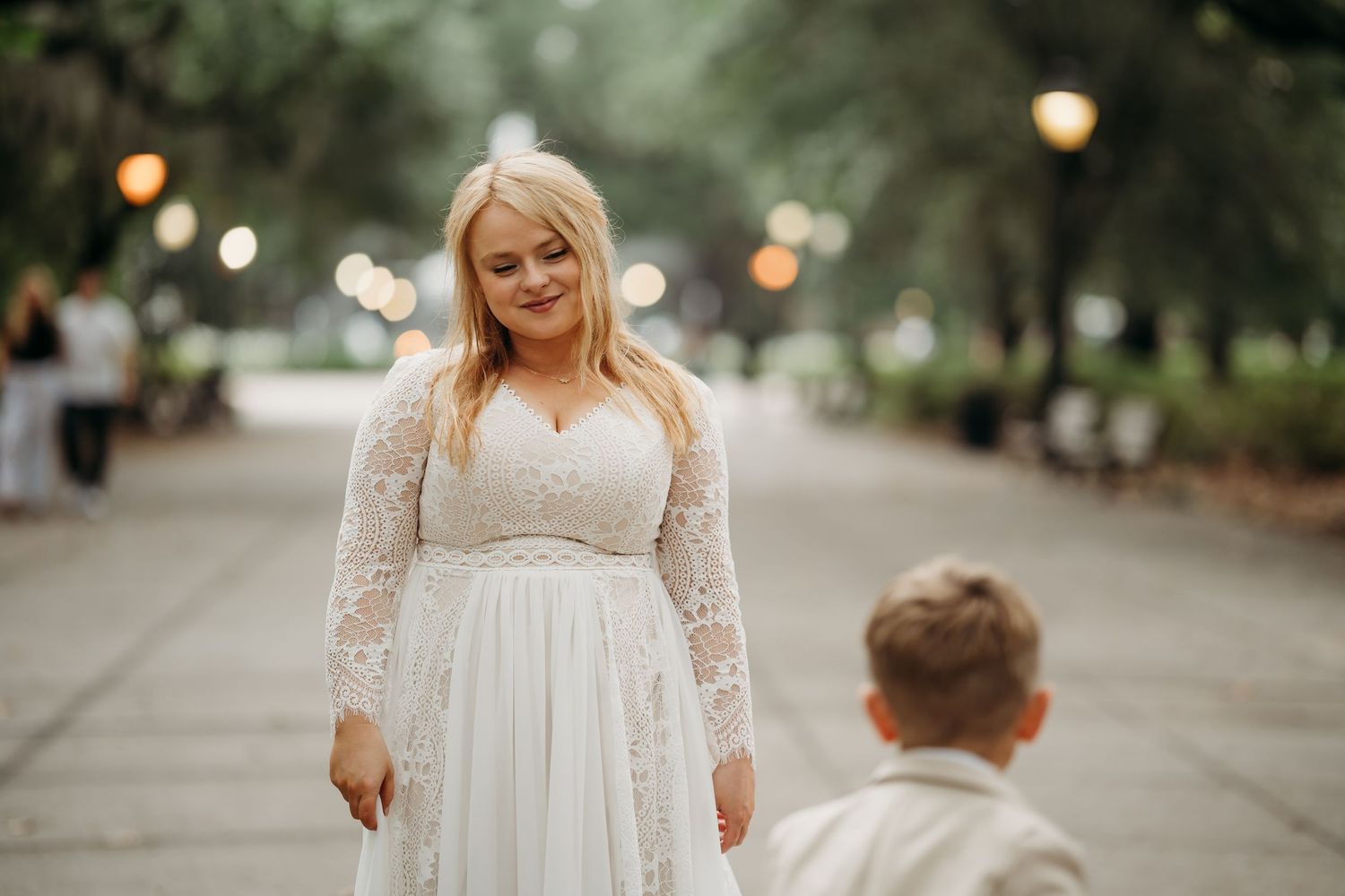 Bride in lace wedding gown holds hands with young boy on bokeh-lit Forsyth Park pathway.