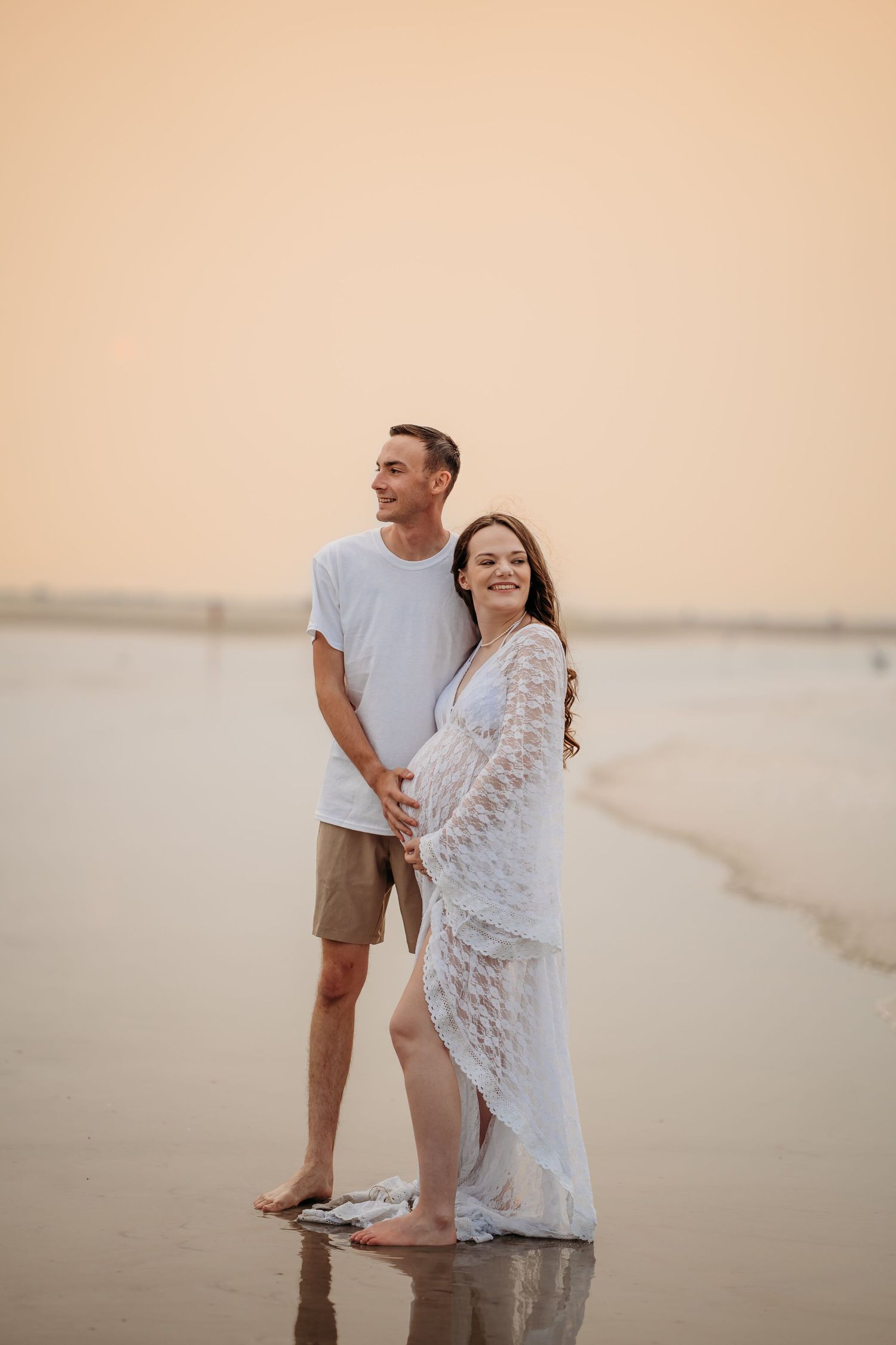 Happy couple embracing on wet beach shoreline at golden sunset, woman in white lace gown.