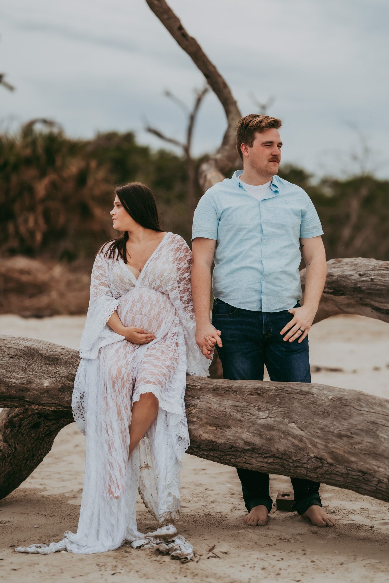 Pregnant couple holding hands leaning on driftwood logs on sandy beach.