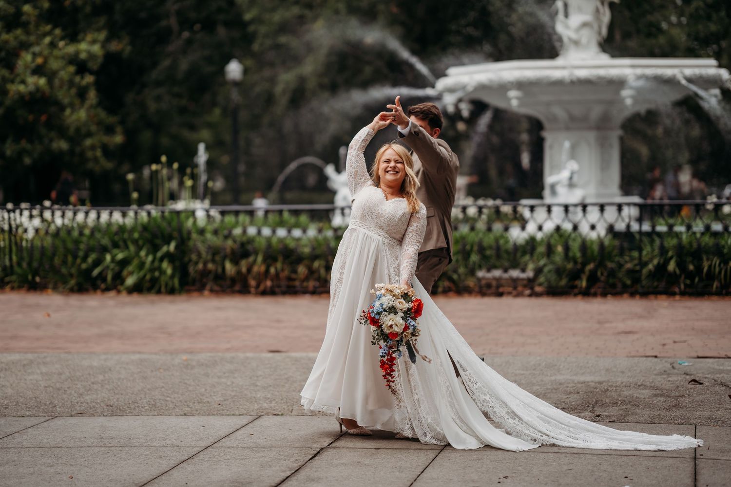 Radiant bride twirls alone in a flowing lace gown holding a colorful bouquet in front of a grand white fountain.