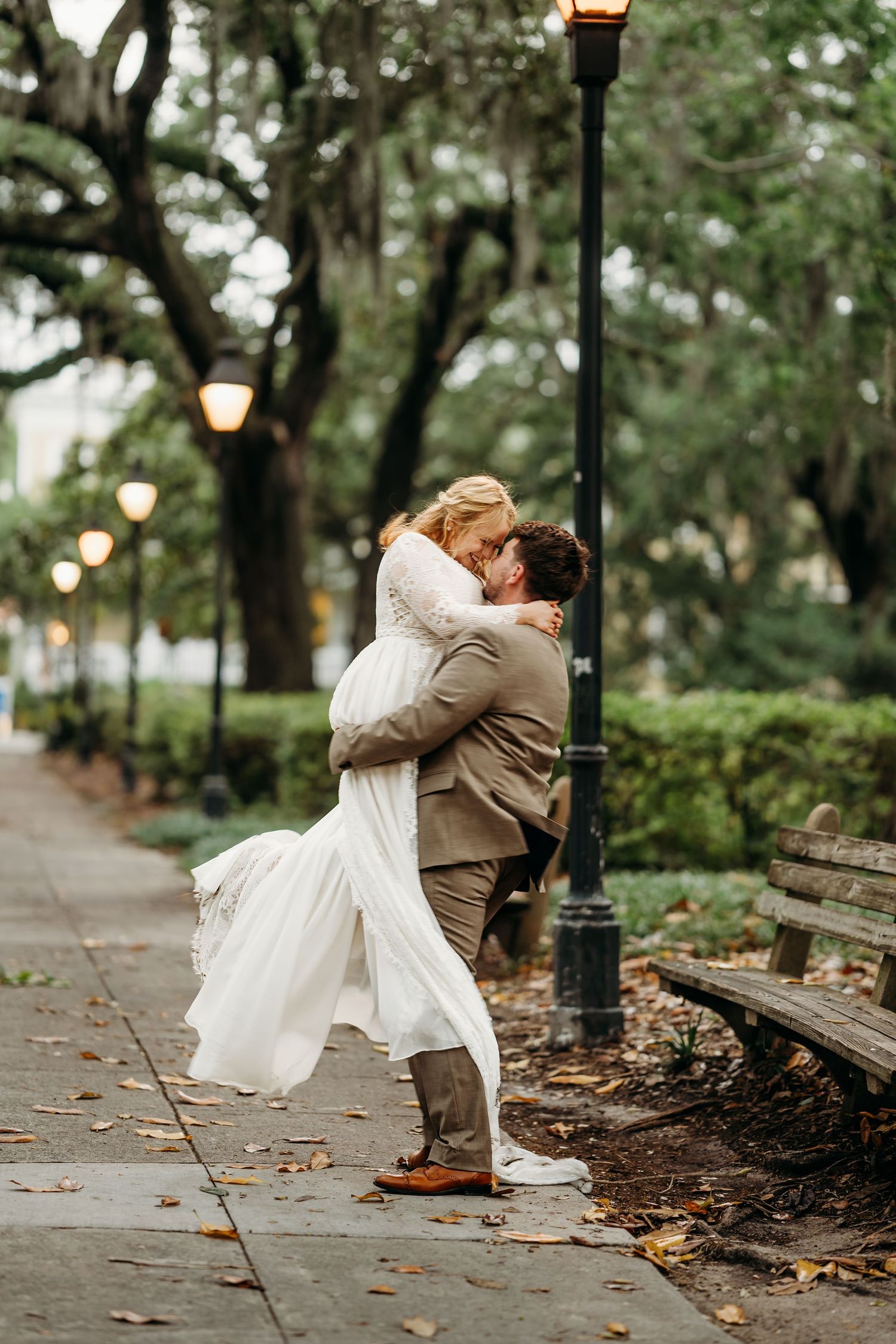 Groom lifts bride in a romantic embrace beside a glowing lamppost on a leaf-covered walkway.
