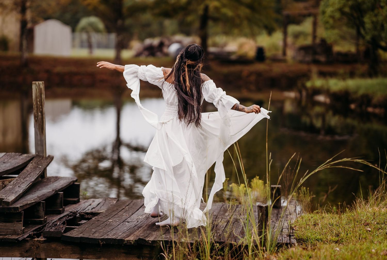 Woman in white billowing dress poses gracefully on aged wooden dock near calm lake.