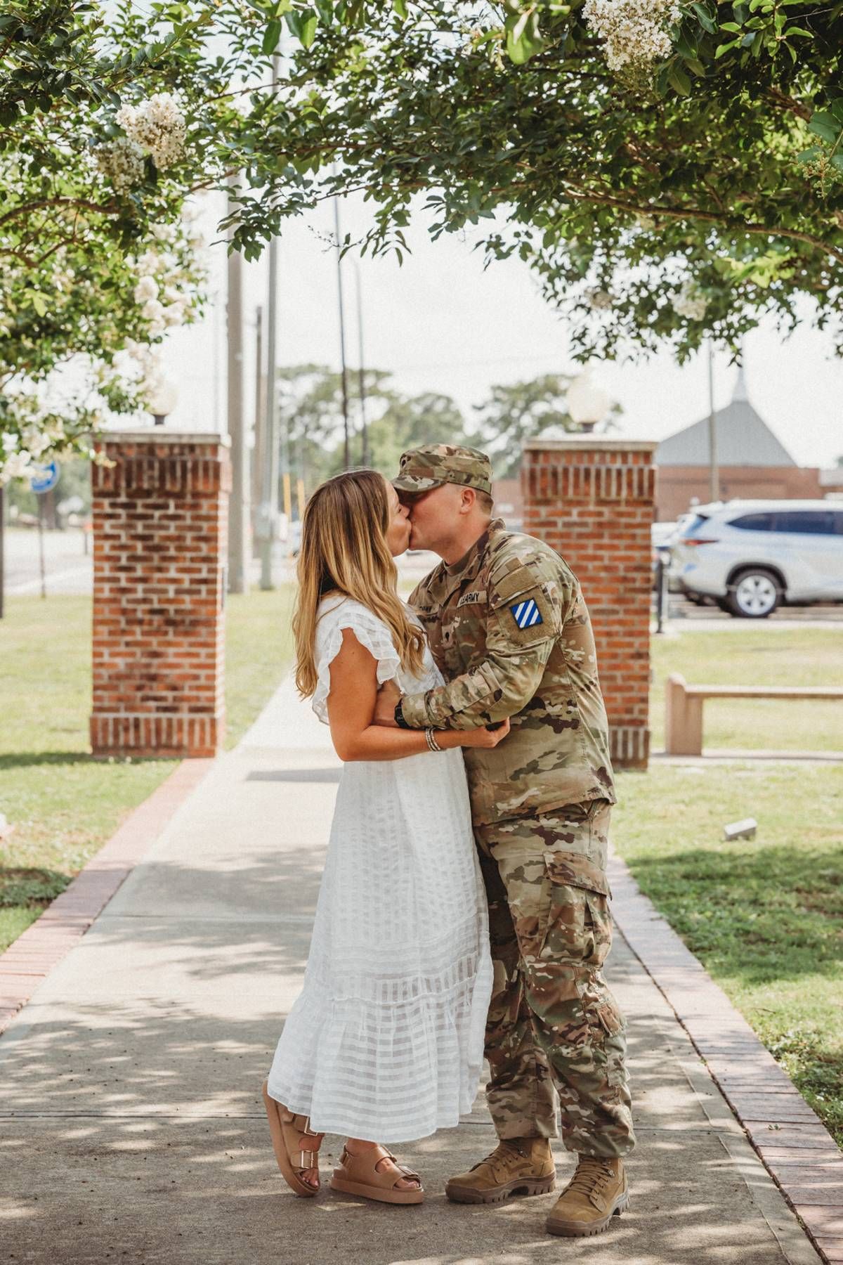 A soldier in camouflage uniform kisses a woman in a white dress on a brick-lined walkway outdoors.