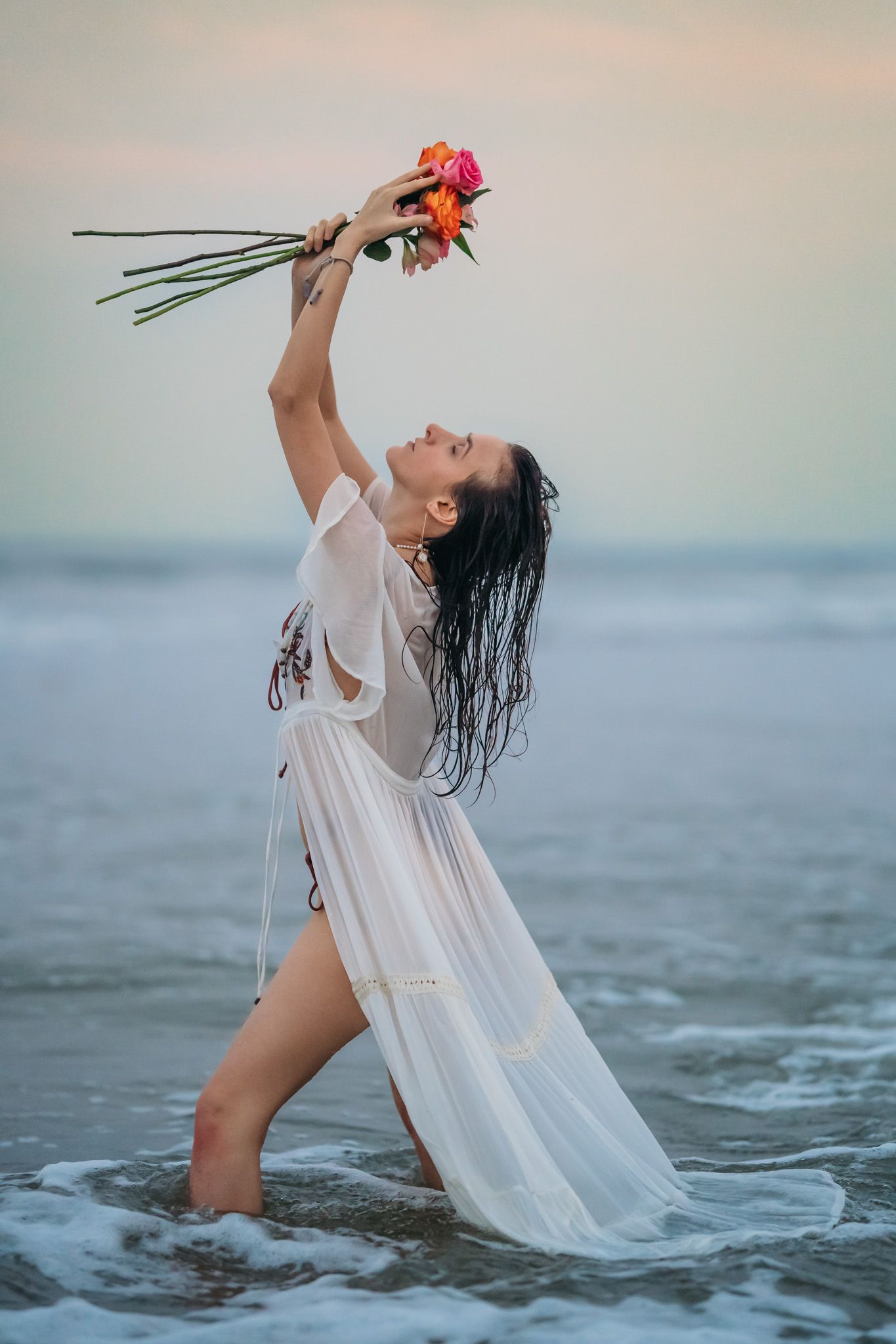 Woman in sheer white dress stands in ocean waves dramatically lifting colorful flowers skyward.